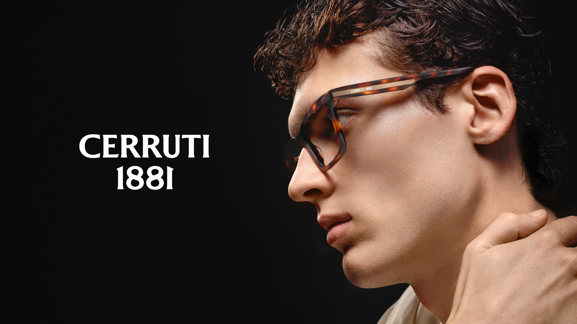 Ein Mann mit einer großen Schildpattbrille blickt vor einem dunklen Hintergrund zur Seite, neben ihm prangt deutlich der Text „CERRUTI 1881“.