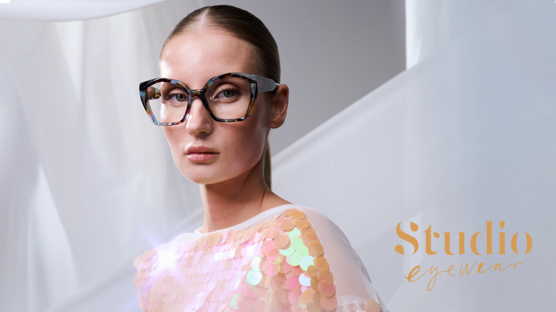 A woman with slicked-back hair wears large, stylish tortoiseshell glasses and a shimmery sequin top. The background is minimal and bright. The text Studio eyewear appears in the lower right corner.