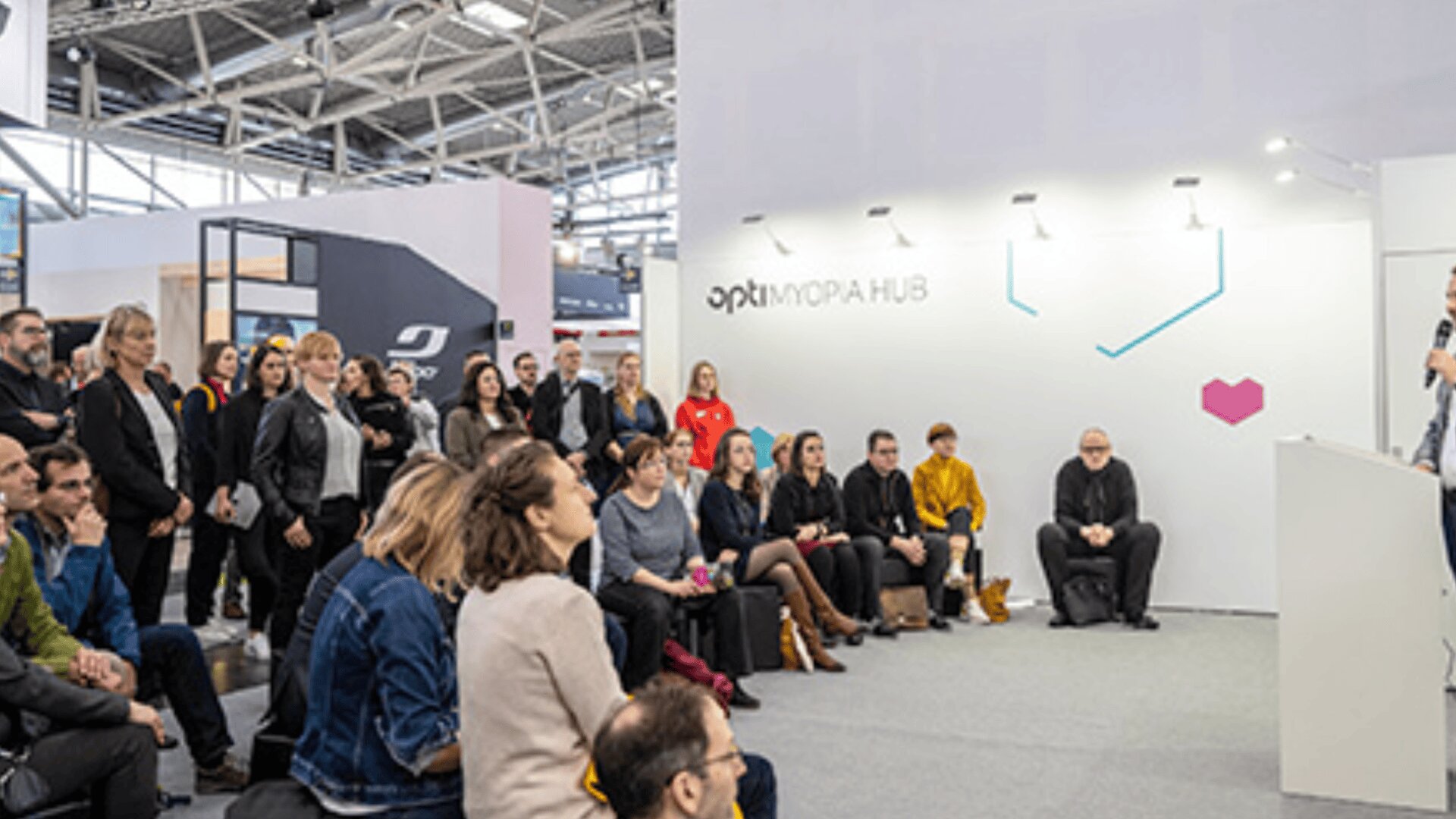 Eine Gruppe von Menschen sitzt und steht aufmerksam vor einem Redner in einem modernen Veranstaltungsraum mit weißem Hintergrund, auf dem "optix MORA HUB" steht.