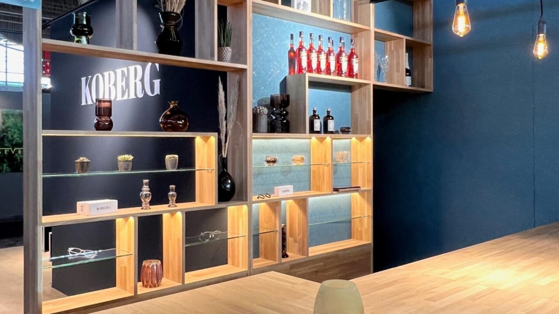 A modern wooden shelving unit with glass shelves, bottles, glassware, and decorative items is displayed in a well-lit room. The word KOBERG is illuminated on a dark wall in the background.