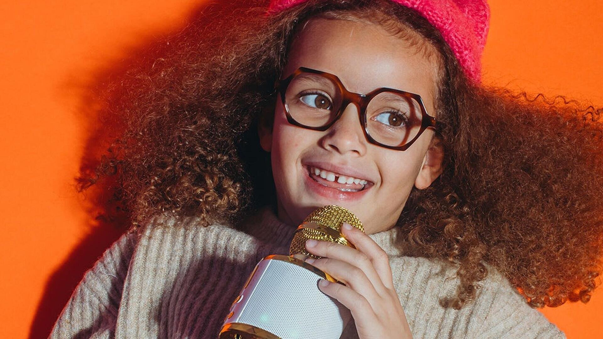 Smiling child with curly hair, wearing glasses and a pink hat, holds a gold and white microphone against an orange background.