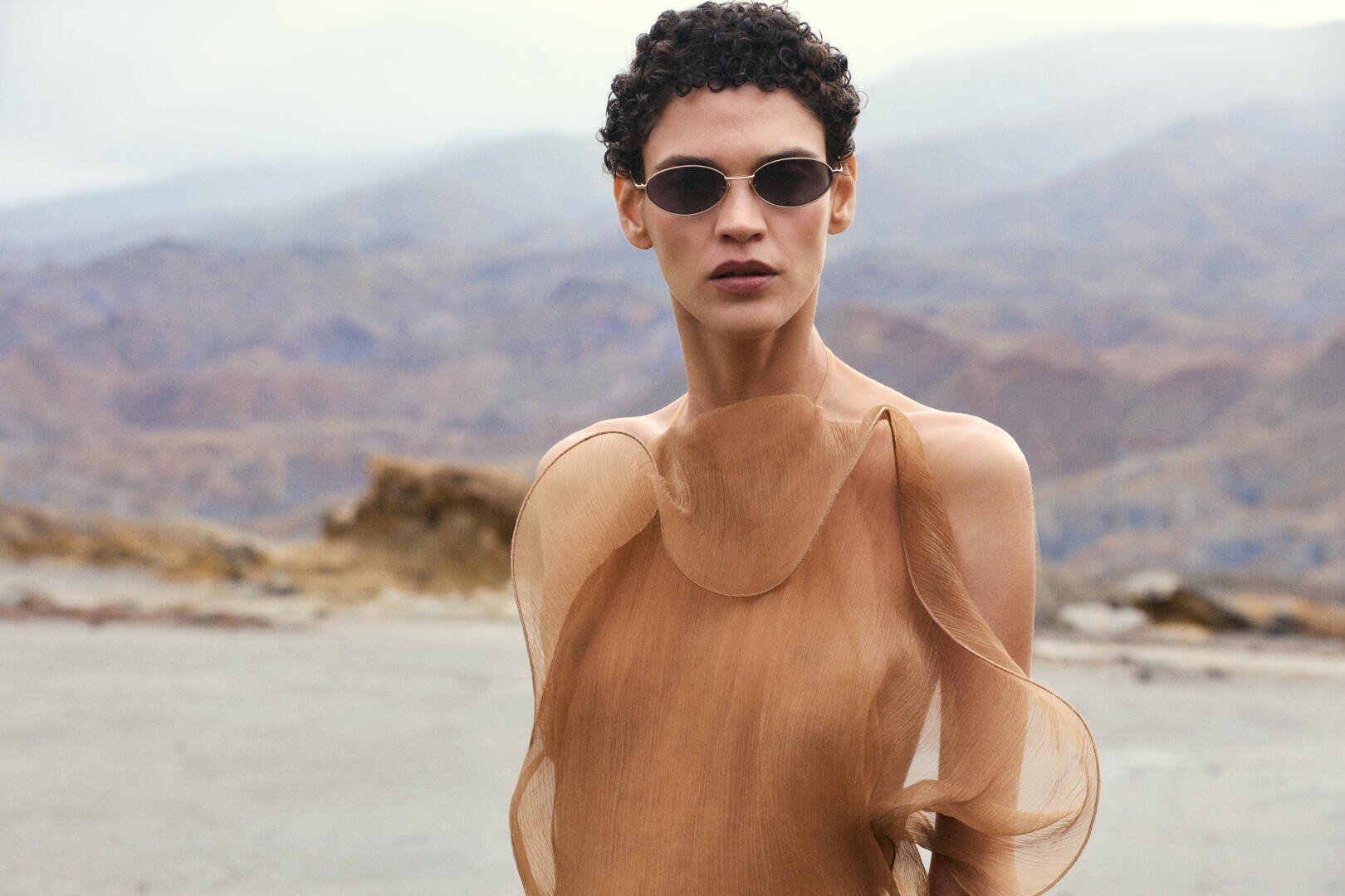 Eine Person mit kurzen lockigen Haaren, die eine Sonnenbrille und ein durchsichtiges, fließendes, hellbraunes Kleid trägt, steht im Freien mit Bergen und wolkenverhangenem Himmel im Hintergrund.