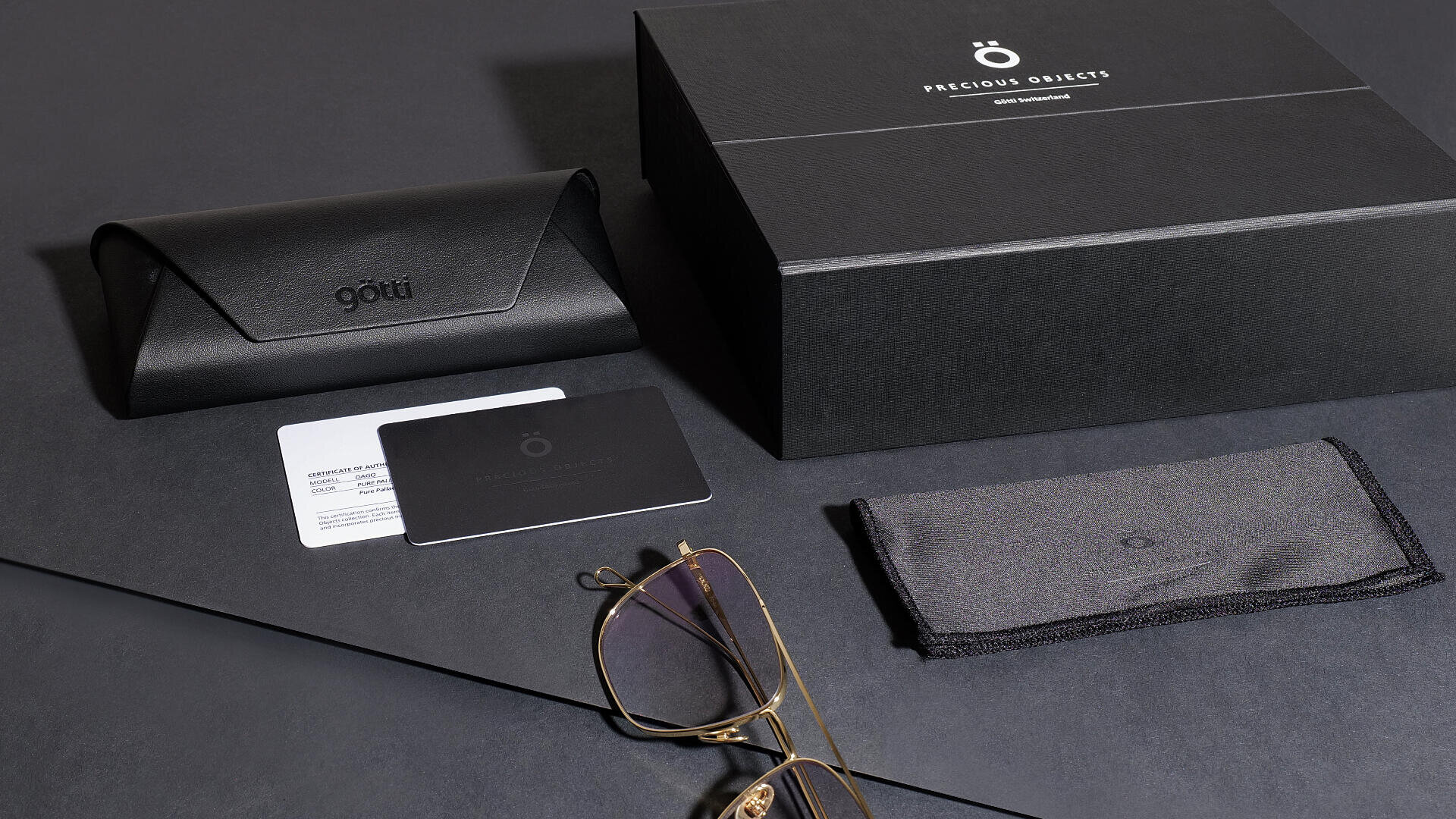 Eine Brille mit Goldrand, ein schwarzes Brillenetui, zwei Karten, ein graues Putztuch und eine große schwarze Schachtel mit der Aufschrift „Precious Objects“ sind ordentlich auf einer dunklen Oberfläche angeordnet.