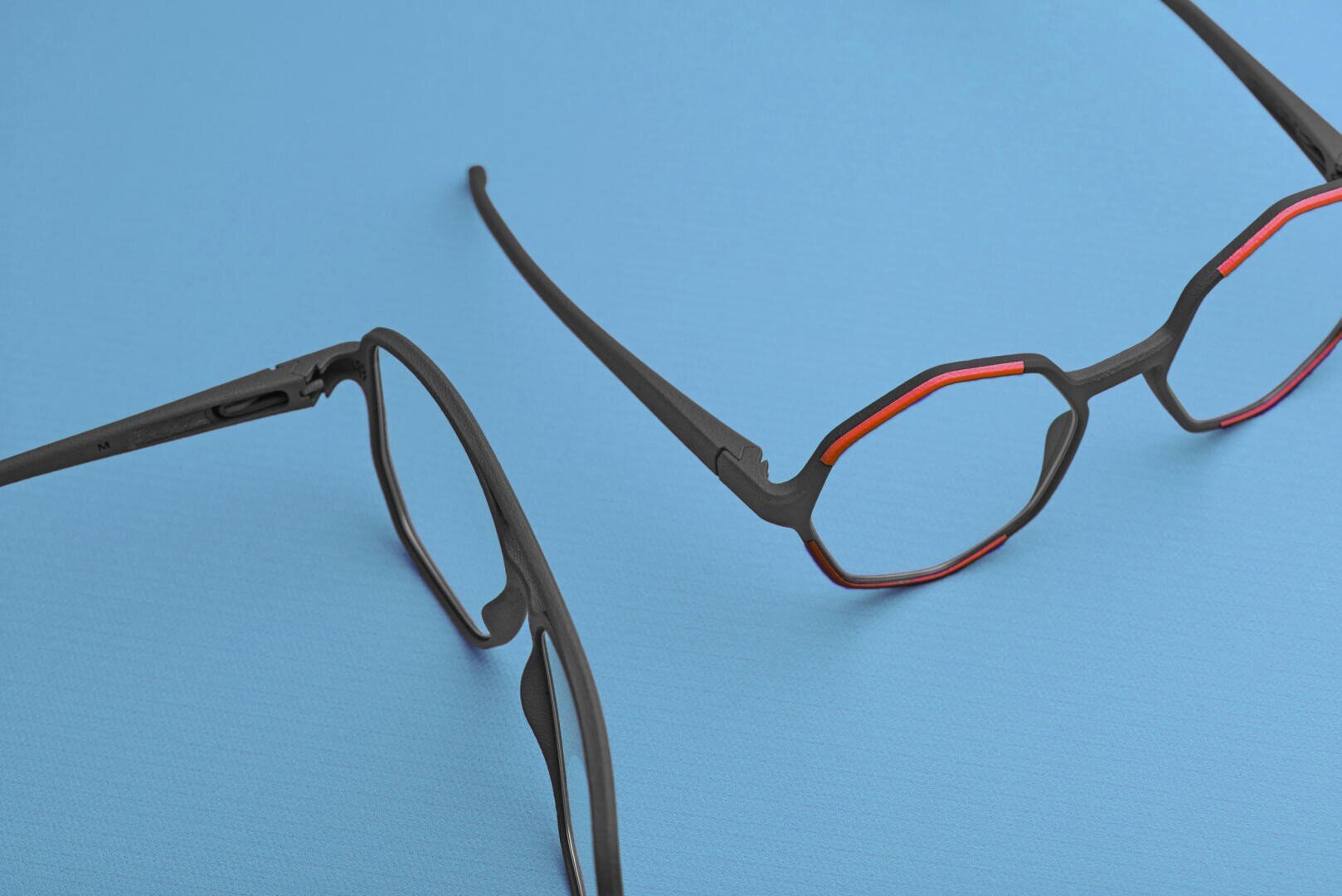 Zwei schwarze Brillen mit geometrischen Rahmen und roten Akzenten sind auf einer flachen, hellblauen Oberfläche platziert. Eine Brille ist vollständig sichtbar, während die andere nur teilweise von der Seite zu sehen ist.