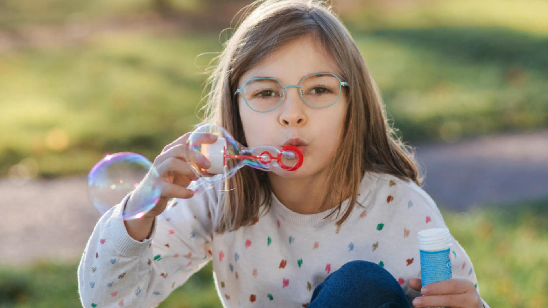 A young girl with glasses and a patterned sweater is blowing bubbles outdoors, holding a bubble wand and a blue bottle, with green grass in the background.