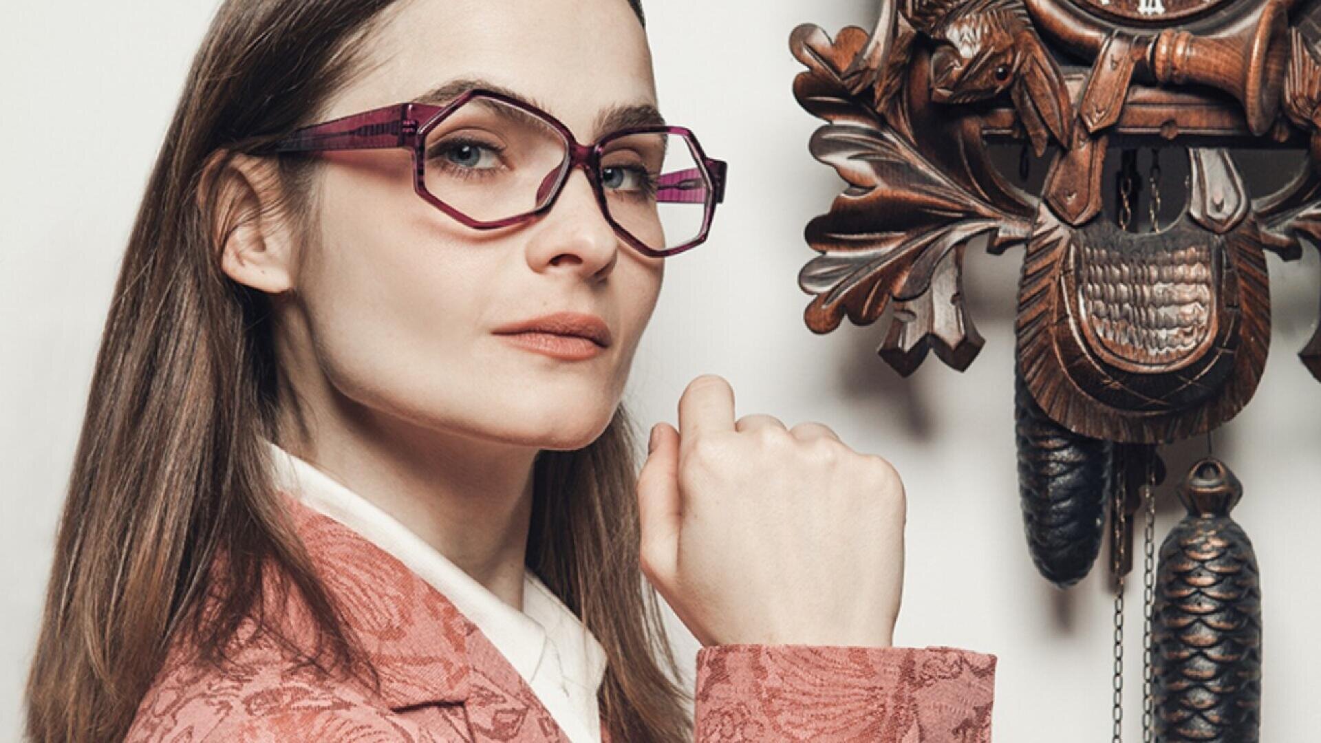 Woman wearing pink geometric glasses and a pink patterned blazer poses confidently with her fist raised near her chin, standing next to an ornate wooden cuckoo clock on a white background.
