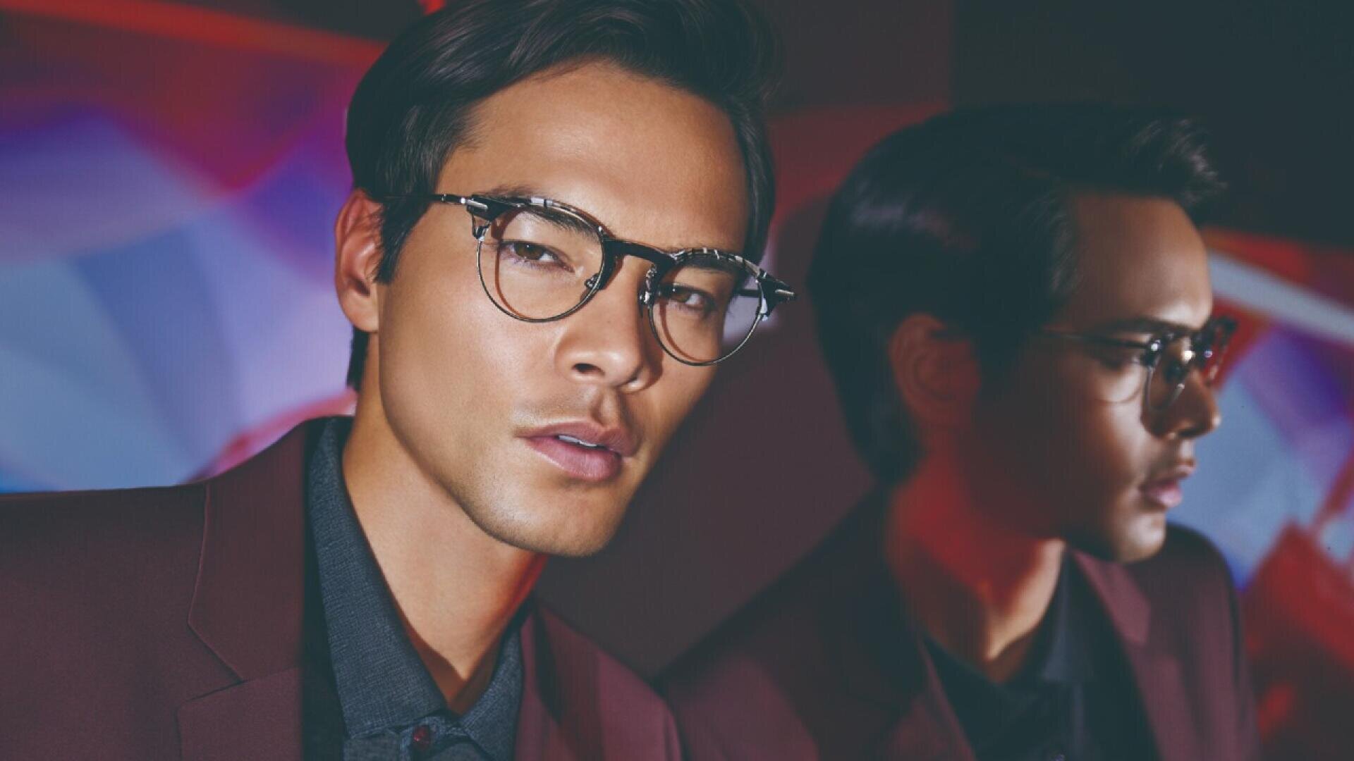 A man with dark hair and glasses wearing a burgundy jacket looks at the camera, while his reflection appears beside him against a blurred, colorful background.