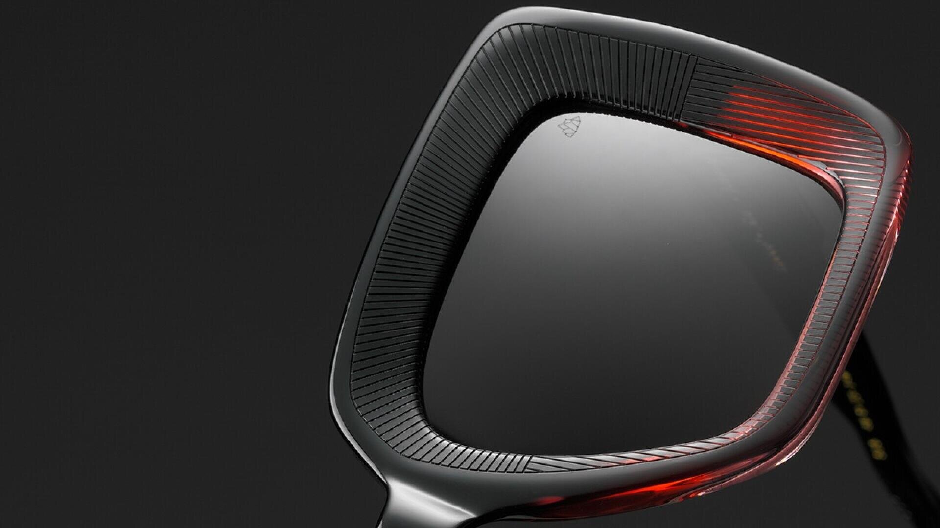Close-up of a modern, square-shaped sunglass lens with a black and red detailed frame against a dark background.