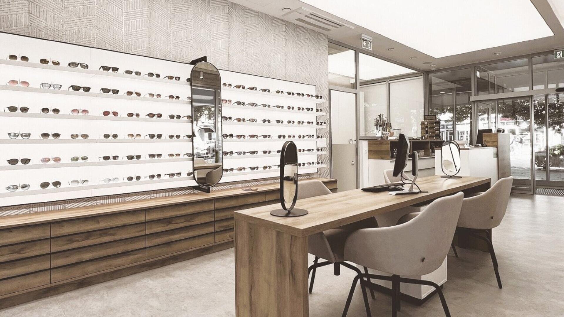 A modern optical store interior featuring a wall display of various eyeglasses and sunglasses, a wooden counter with two chairs, tabletop mirrors, and large windows letting in natural light.