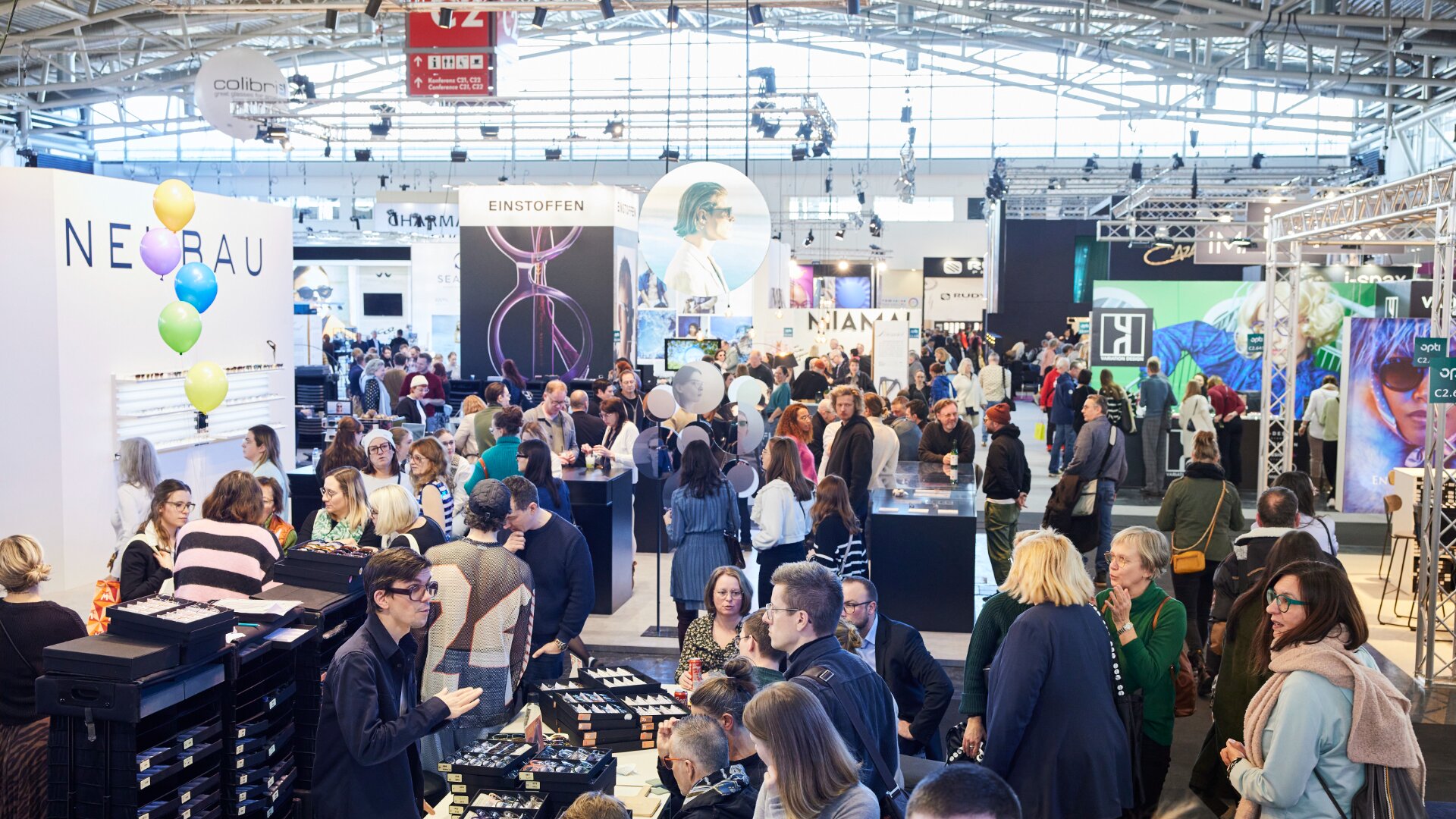 A busy indoor trade show with many people walking, talking, and browsing booths displaying eyewear products. Colorful balloons and branded signs are visible among the lively crowd in the exhibition hall.