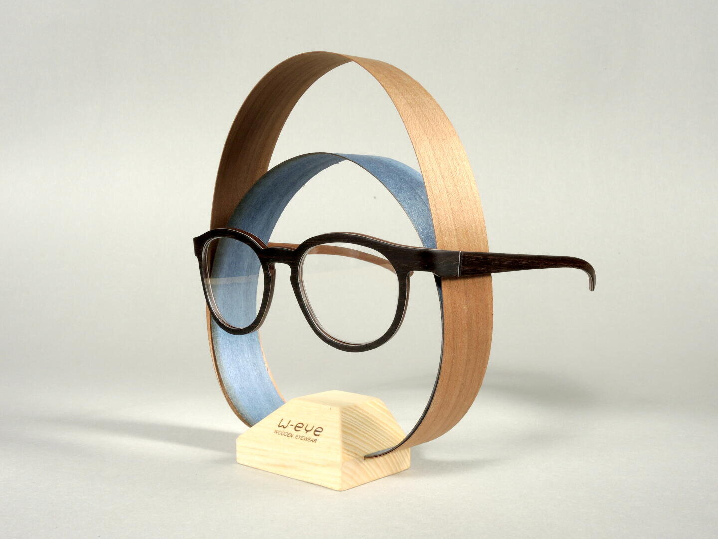 A pair of wooden eyeglasses is displayed on a light wooden stand, with two curved wooden veneers forming oval shapes around the glasses on a plain light background.