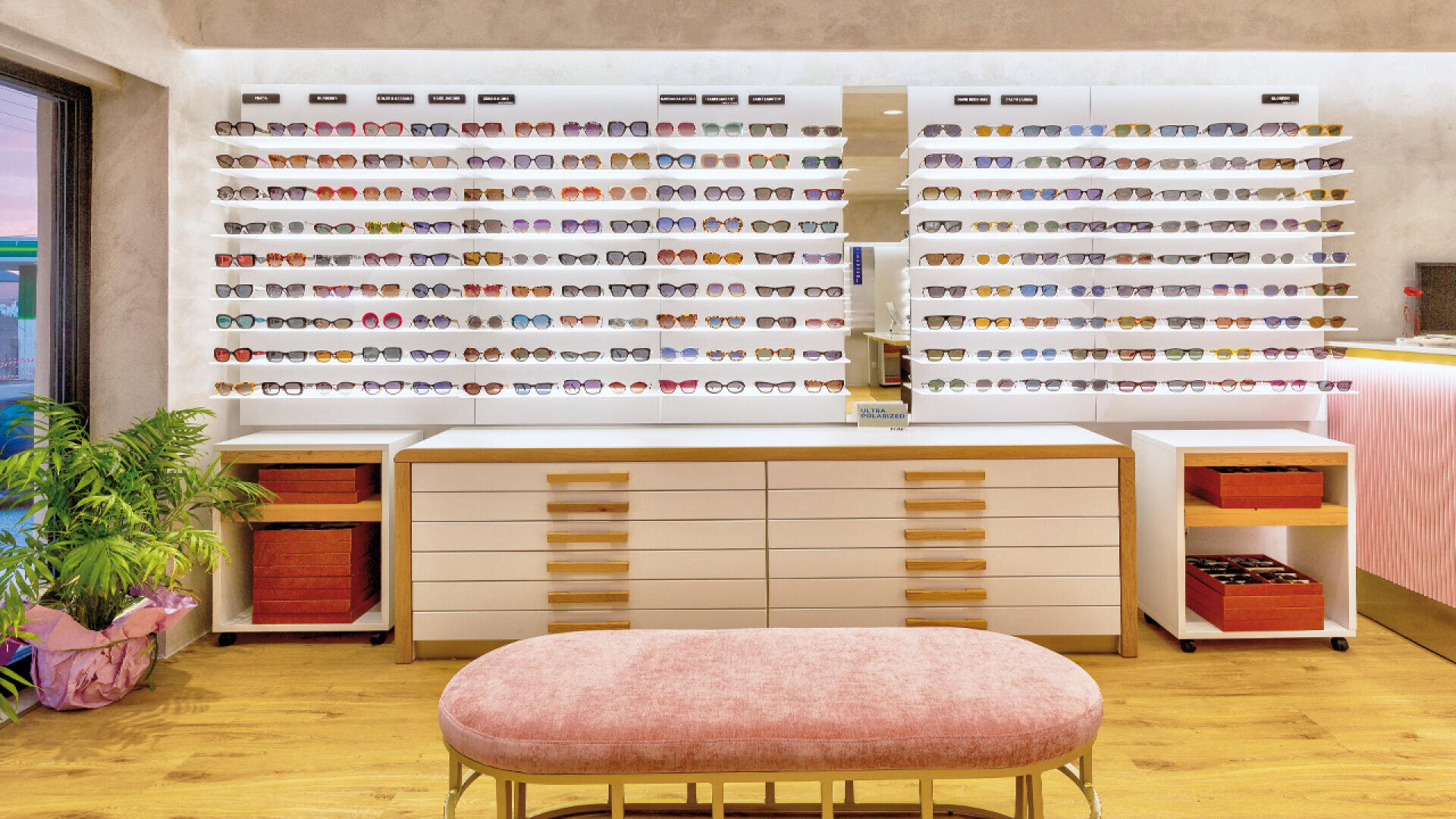 Ein modernes Brillengeschäft mit weißen Schubladen und Regalen, in denen zahlreiche Brillen und Sonnenbrillen ausgestellt sind. Im Vordergrund steht eine rosa gepolsterte Bank, links davon eine grüne Topfpflanze.