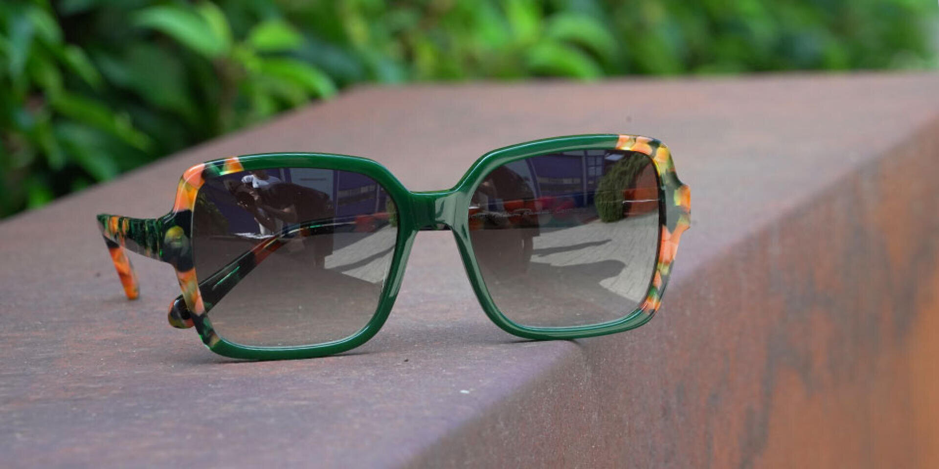 Eine übergroße Sonnenbrille mit grün-orange marmoriertem Gestell liegt auf einem braunen Sims, während das grüne Laub im Hintergrund verschwimmt.