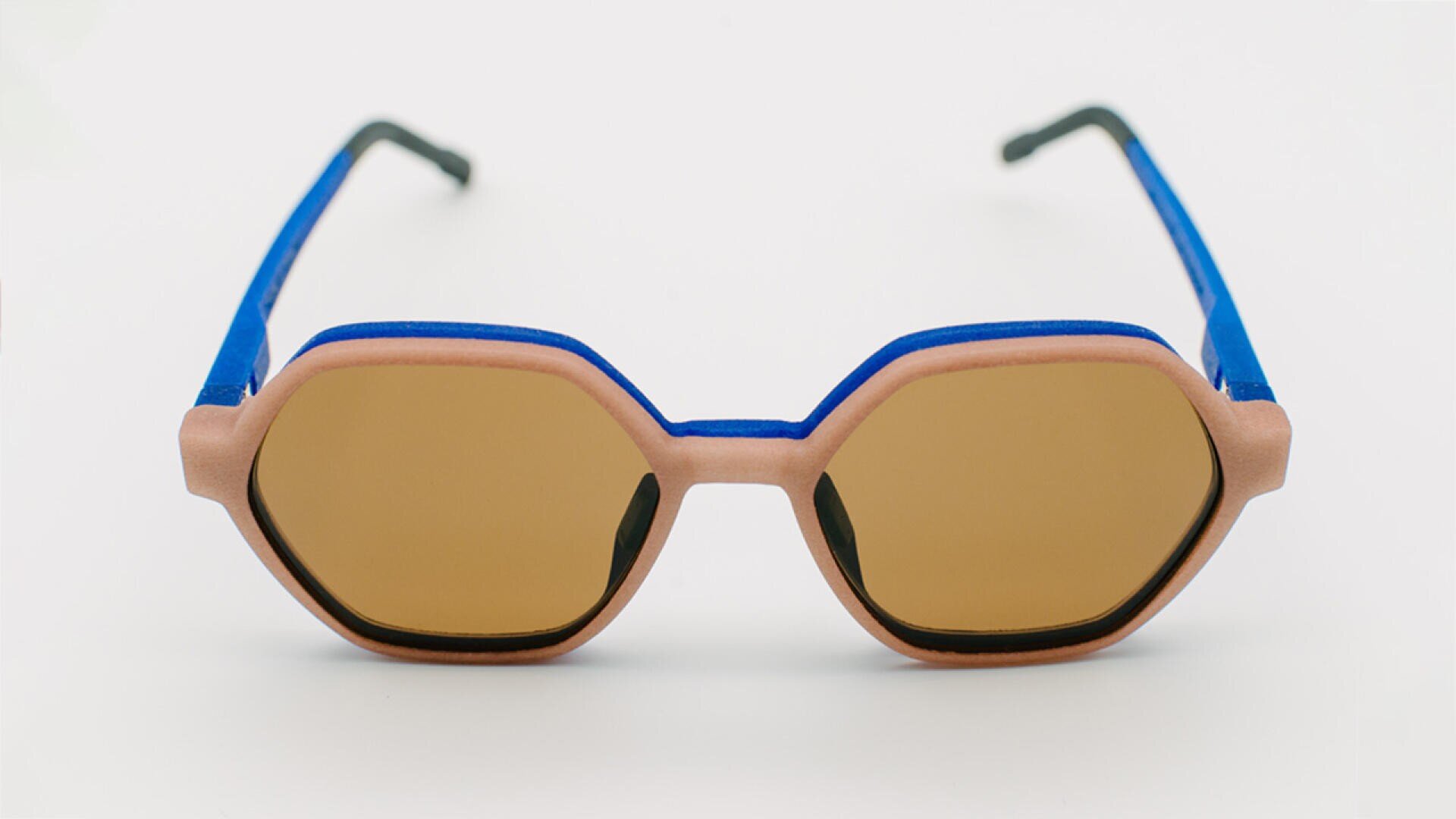 Eine achteckige Sonnenbrille mit hellbraunen Gläsern, rosafarbener Rahmenfront und blauen Bügeln mit schwarzen Spitzen, abgebildet auf weißem Hintergrund.