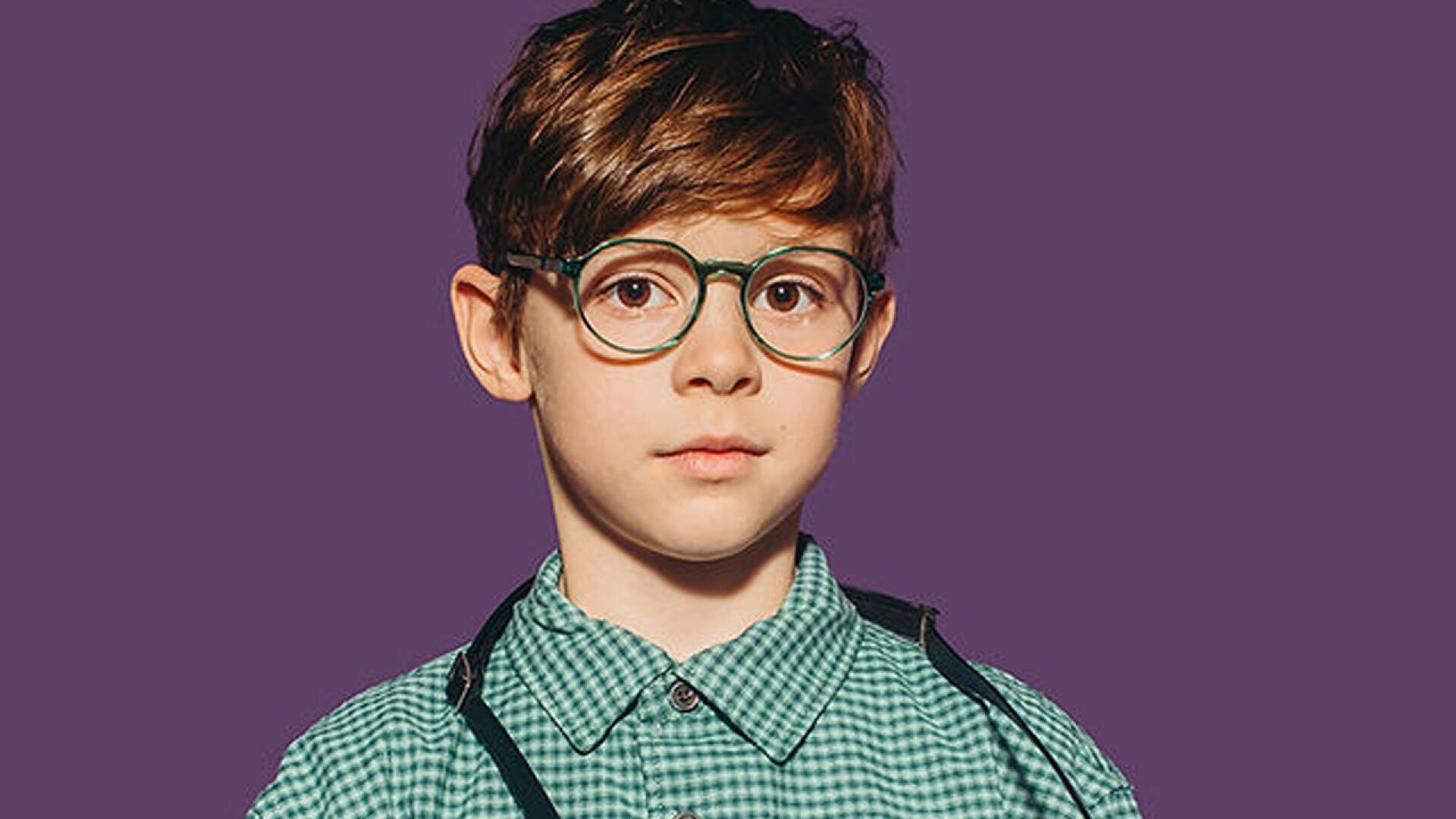Ein kleiner Junge mit hellbraunem Haar, einer runden Brille, einem grün karierten Hemd und schwarzen Hosenträgern steht vor einem einfarbig violetten Hintergrund.