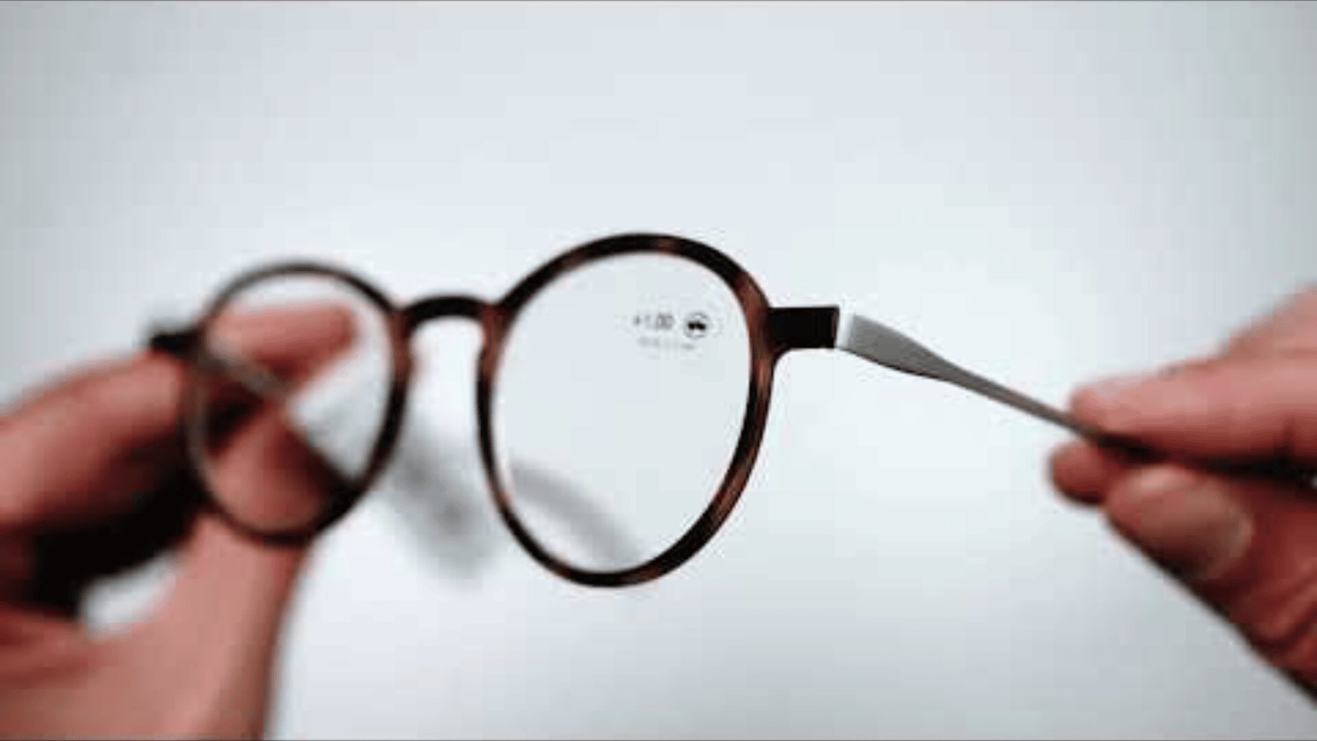 Eine Nahaufnahme von zwei Händen, die eine runde Brille mit braunem Rahmen vor weißem Hintergrund halten. Der Fokus liegt auf der Brille, auf einem Glas ist ein kleiner Aufkleber mit Angaben zur Sehstärke zu sehen.
