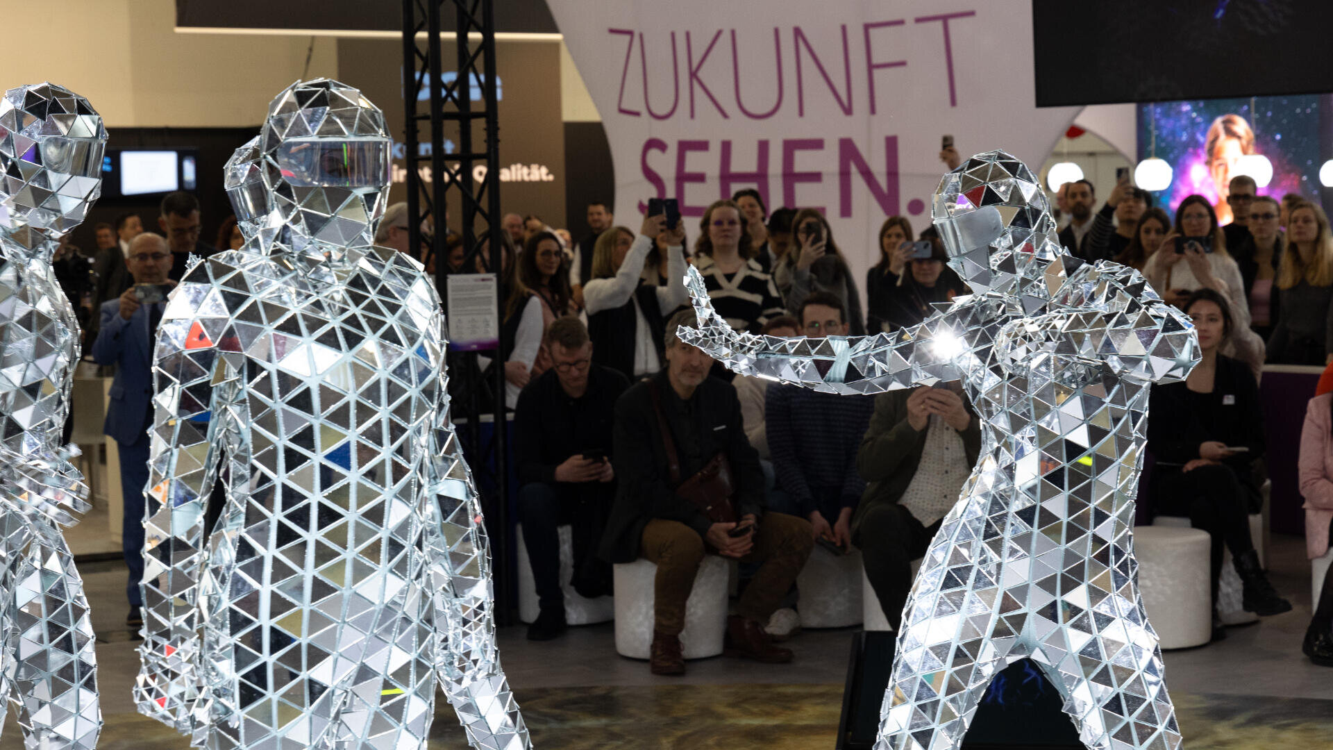 Drei Personen in reflektierenden Anzügen treten bei einer Indoor-Veranstaltung vor einem sitzenden Publikum auf. Im Hintergrund steht auf einem großen Schild „ZUKUNFT SEHEN“.