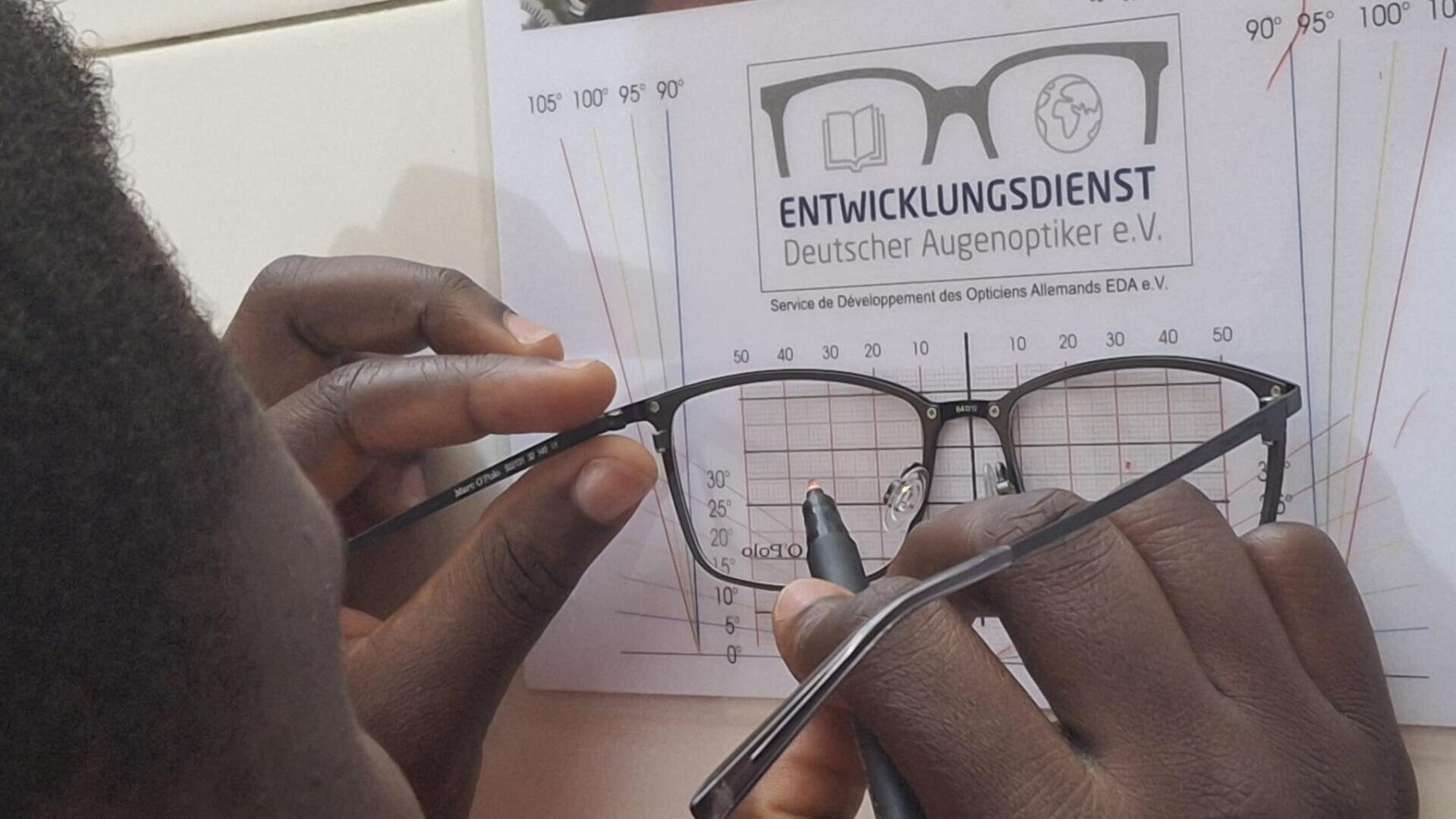 Eine Person misst Brillengestelle mit einem Lineal aus und richtet sie nach einer Tabelle mit der Aufschrift ENTWICKLUNGSDIENST Deutscher Augenoptiker e.V." und Winkelmarkierungen aus.
