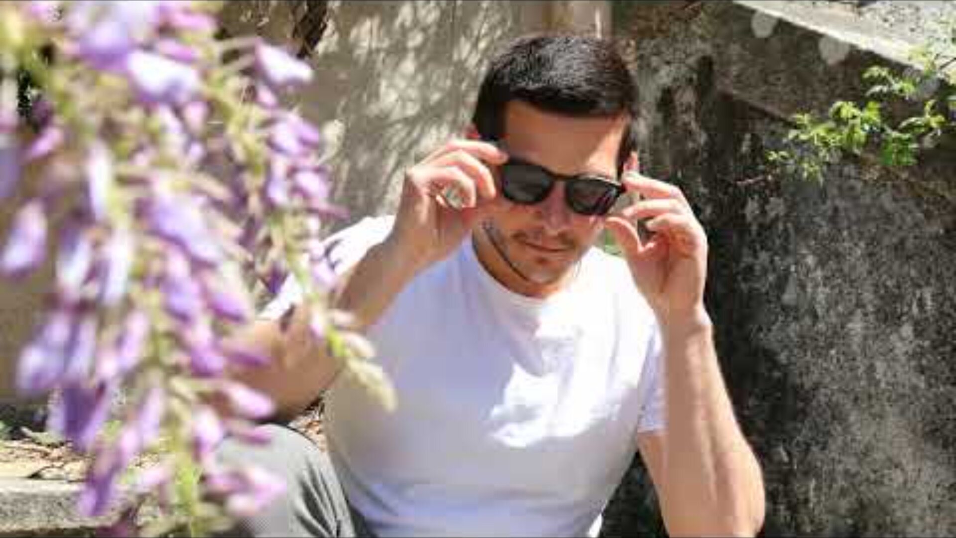 Ein Mann in einem weißen T-Shirt sitzt im Freien auf steinernen Stufen und rückt seine schwarze Sonnenbrille zurecht. Im Vordergrund sind lila Blumen zu sehen, die leicht unscharf sind. Das Sonnenlicht wirft Schatten auf die Szene.