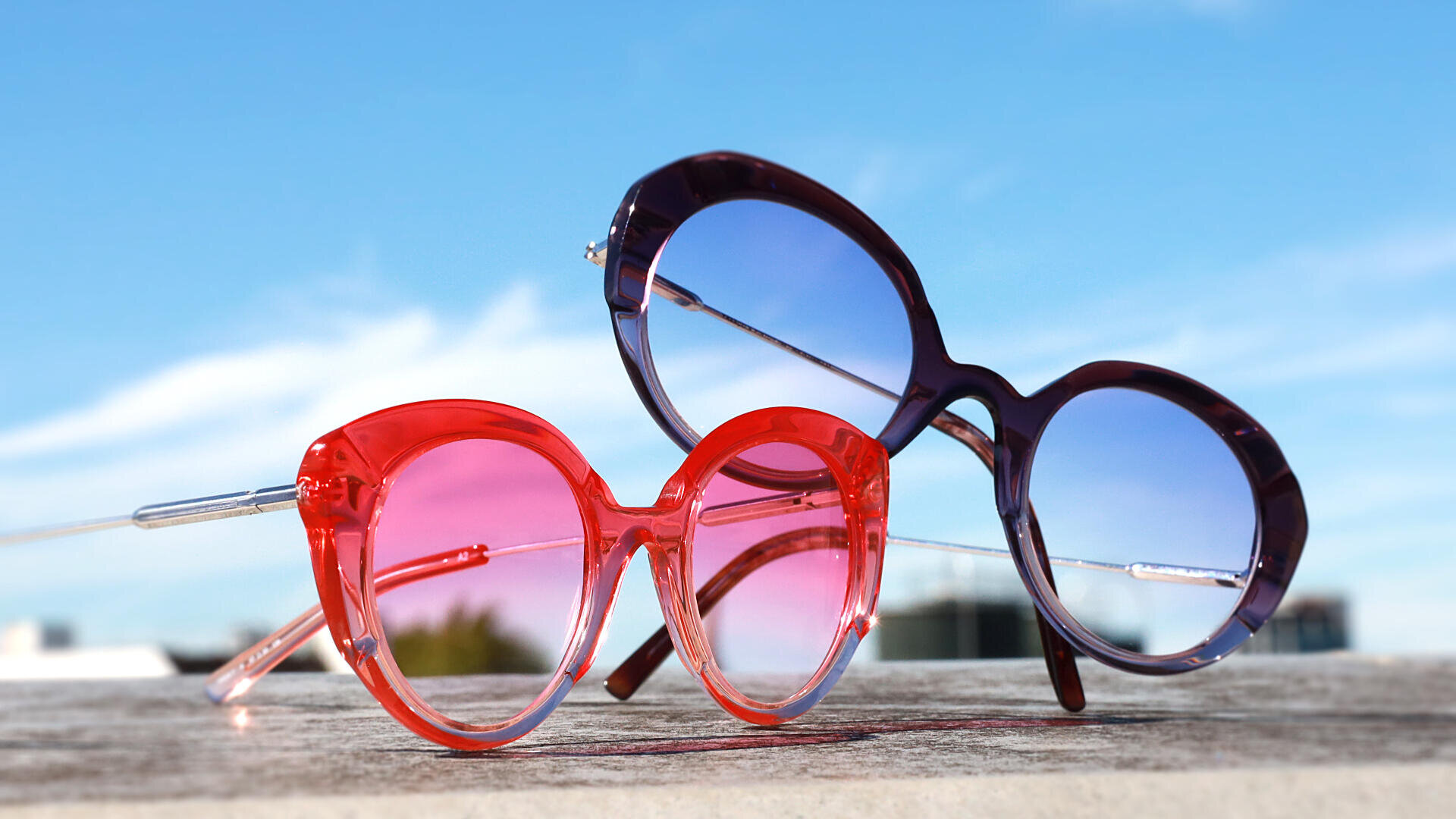 Zwei Paar runde Sonnenbrillen, eine mit rosa Rahmen und Gläsern, die andere mit violettem Rahmen und blauen Gläsern, liegen im Freien auf einer Holzoberfläche mit blauem Himmel und verschwommenen Gebäuden im Hintergrund.