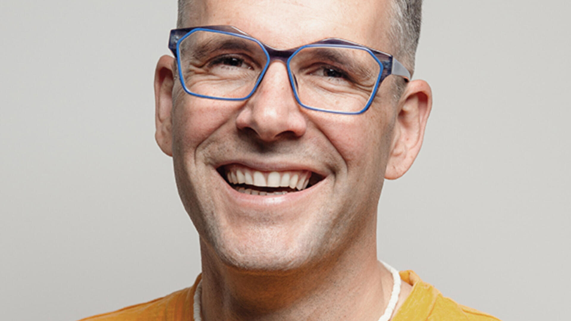 Ein lächelnder Mann mit kurzen Haaren, einer Brille mit blauem Rahmen und einem gelben Hemd blickt vor einem schlichten hellen Hintergrund direkt in die Kamera.