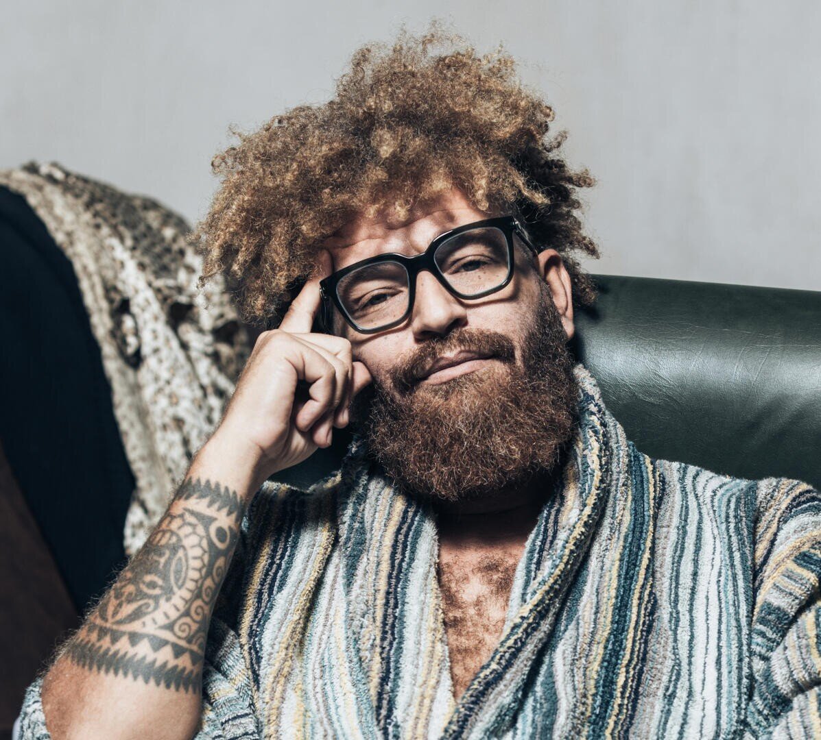 Ein Mann mit lockigem Haar, Vollbart und Brille sitzt auf einem grünen Stuhl. Er trägt ein gestreiftes Gewand und hat einen nachdenklichen Gesichtsausdruck. Eine Hand ruht auf seiner Schläfe und gibt den Blick auf eine Tätowierung auf seinem Unterarm frei.