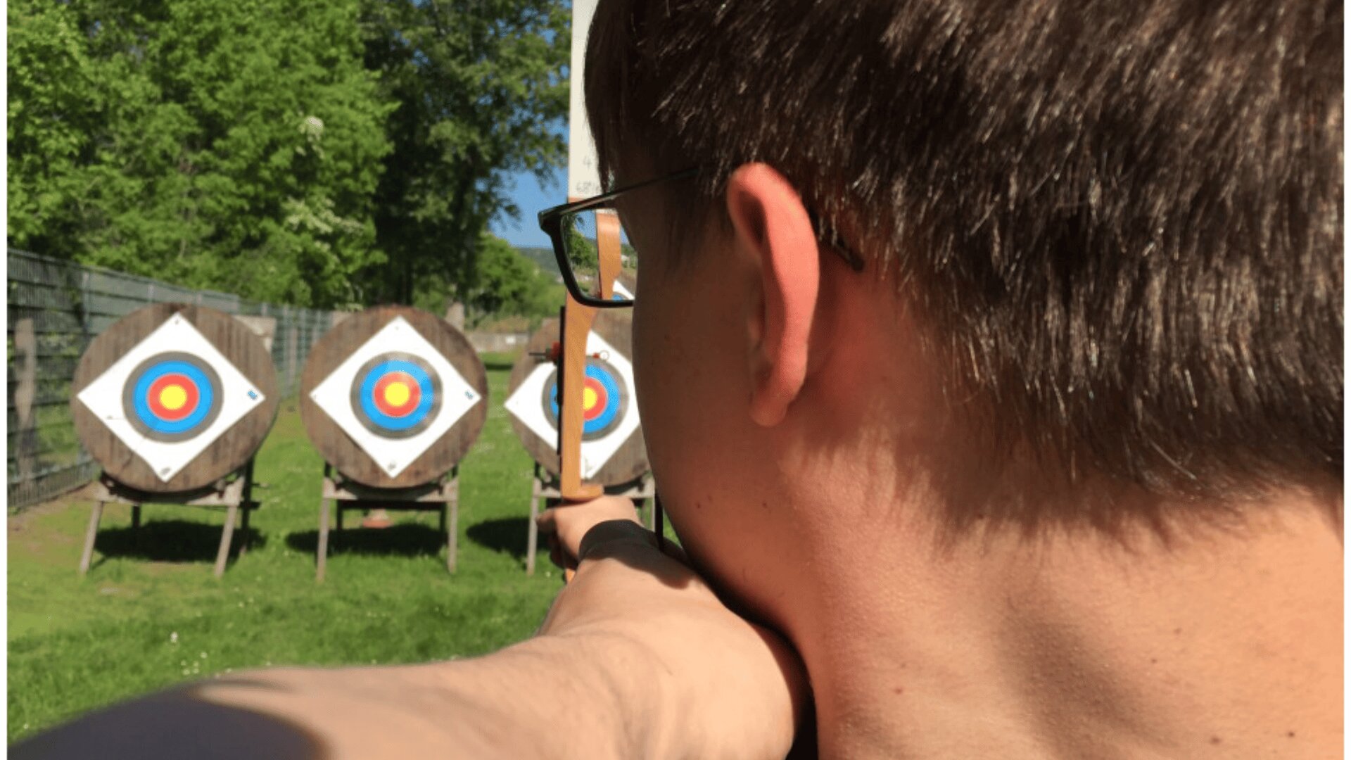 A person with short hair and glasses is aiming a bow at three archery targets placed on a grassy field, seen from behind on a sunny day.