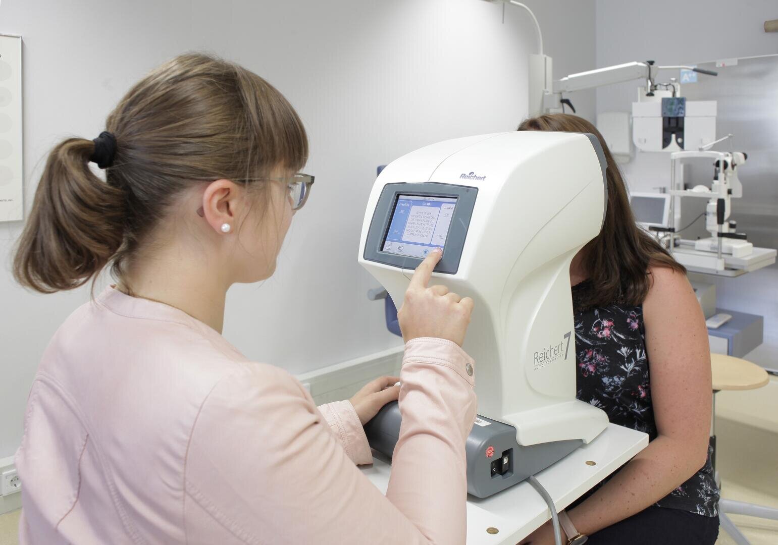 Eine Frau bedient in einer Klinik ein Augenuntersuchungsgerät für eine andere Frau, die davor sitzt. Die Bedienerin interagiert mit dem Touchscreen-Display des Geräts.