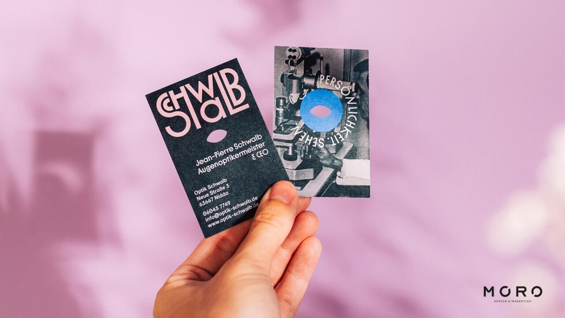 A hand holds two business cards against a blurred pink and purple background. One card is dark with bold text, and the other has a collage-style design with a blue circle. The MGRO logo appears at the bottom right.
