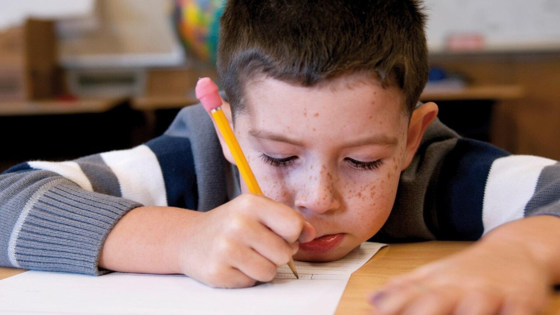 A young boy with freckles, wearing a striped sweater, leans over a desk and concentrates while writing with a pencil on a sheet of paper.