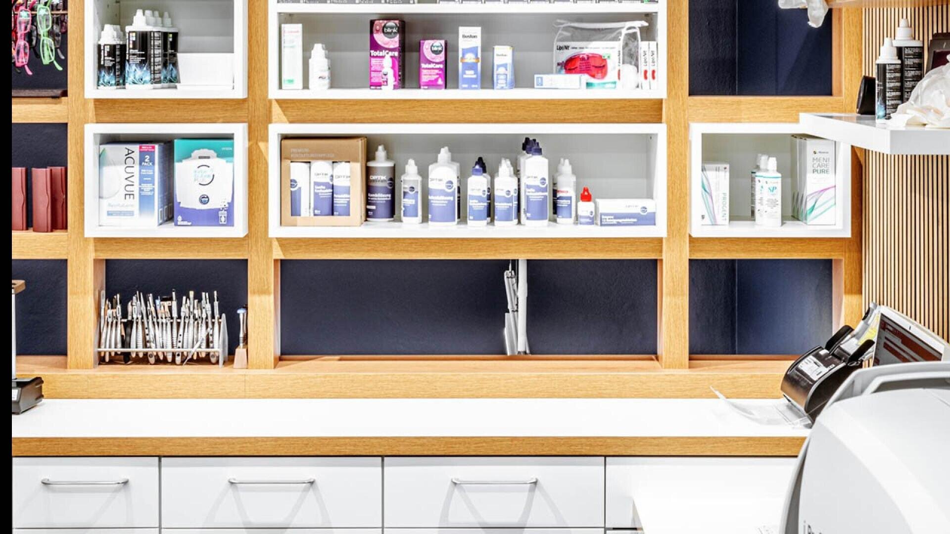 A modern pharmacy or clinic shelf with various medical supplies, bottles, and dental products neatly arranged in wooden cubbies above a white counter with drawers and equipment.