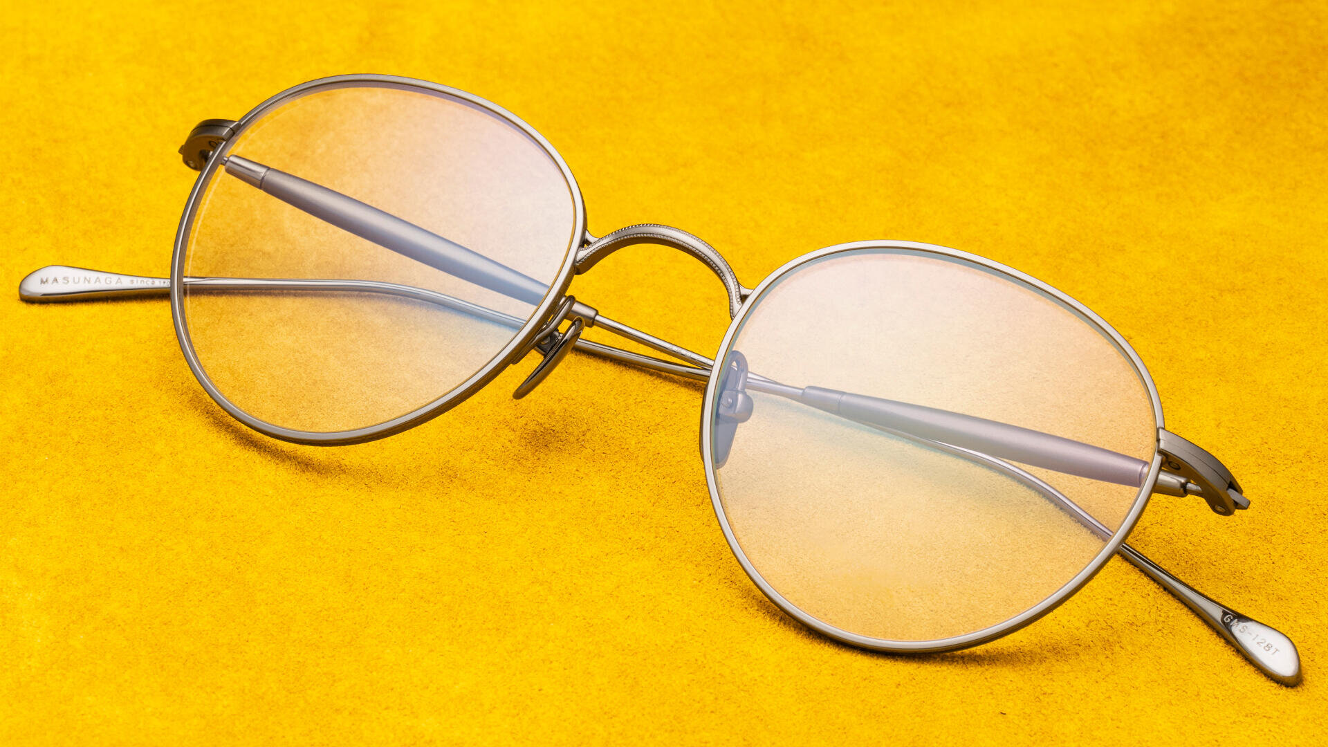 Eine runde Brille mit Metallrahmen und klaren Gläsern liegt auf einer leuchtend gelben Oberfläche, die das Licht leicht auf die Gläser reflektiert.
