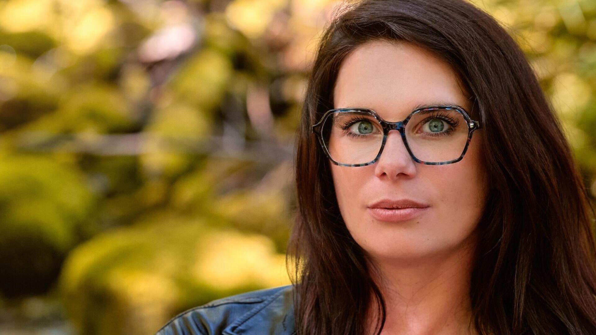 A woman with long brown hair and glasses looks at the camera. She is wearing a black jacket, and the blurred background features green foliage and sunlight.