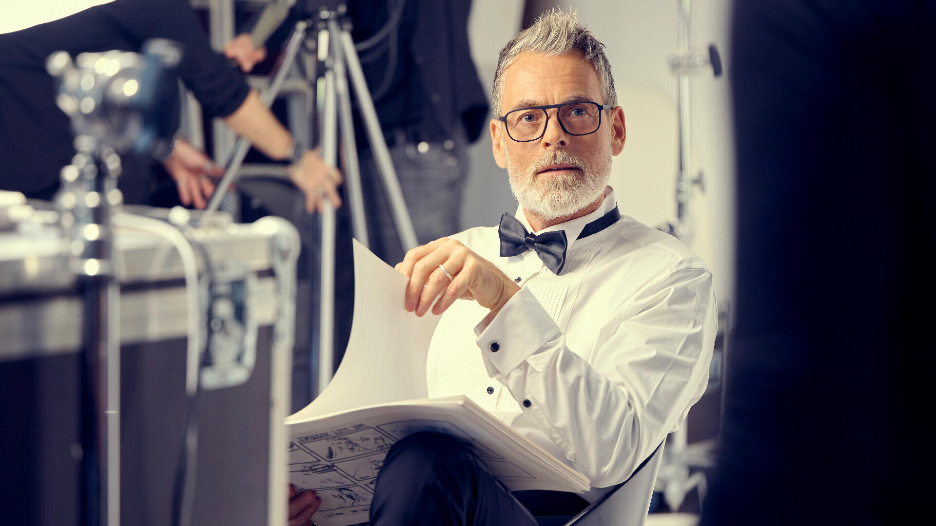 Ein Mann mit grauem Haar und Brille, gekleidet in ein weißes Hemd und eine schwarze Fliege, sitzt mit Papieren in der Hand an einem Filmset. Er wirkt aufmerksam, im Hintergrund sind Kameras und Crew zu sehen.