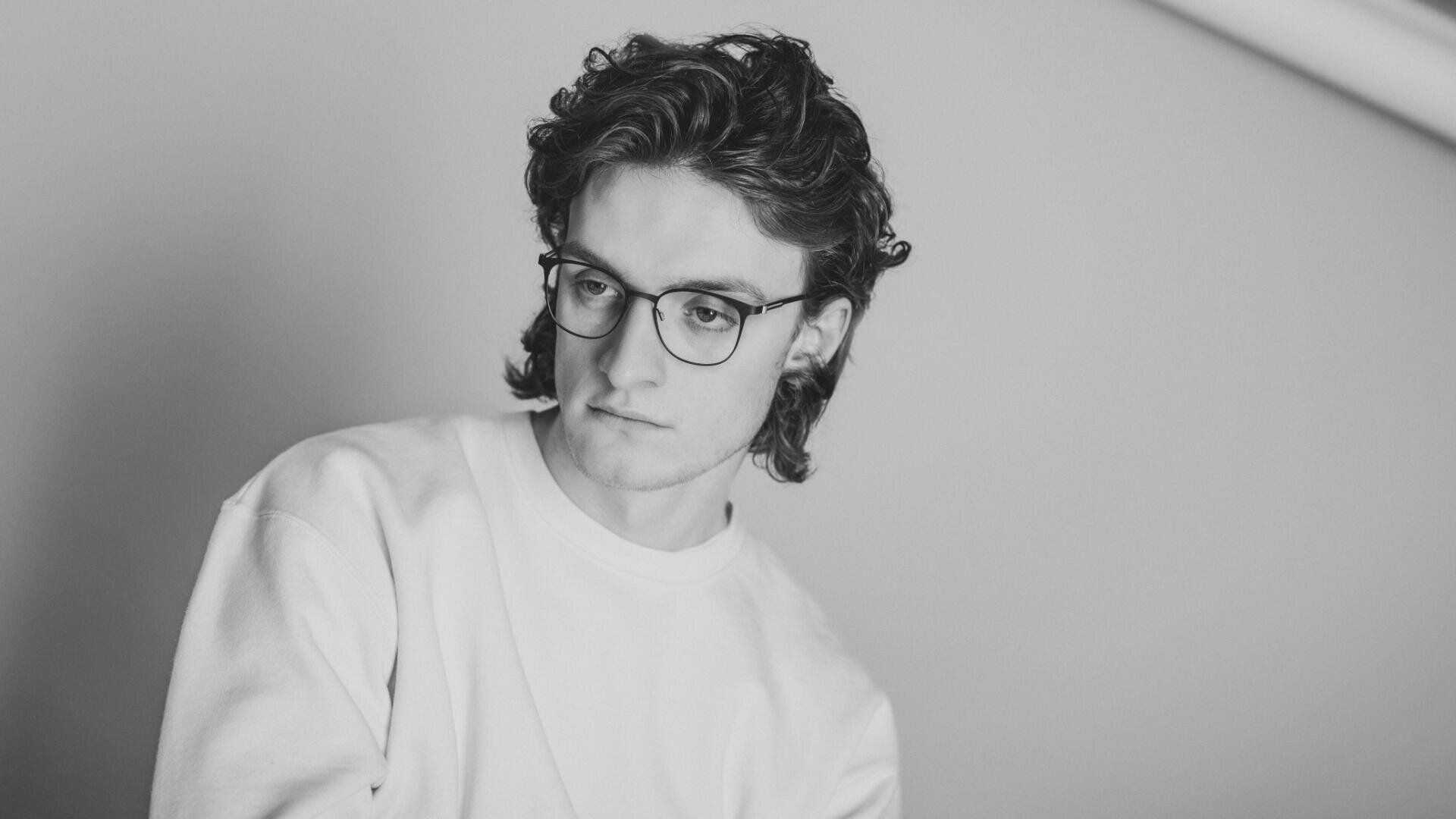 Black and white photo of a young person with wavy hair wearing glasses and a plain sweatshirt, looking down thoughtfully against a plain background.