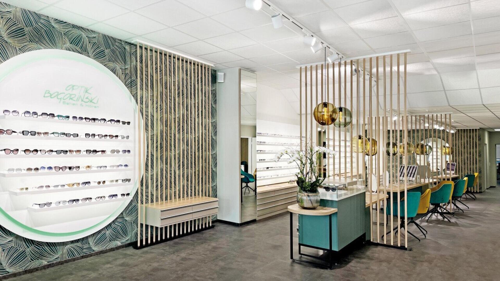 A modern optical store interior with glasses displayed on backlit shelves, wooden slats separating seating areas with yellow chairs, green accents, round hanging lights, and decorative plants. The space is bright and contemporary.