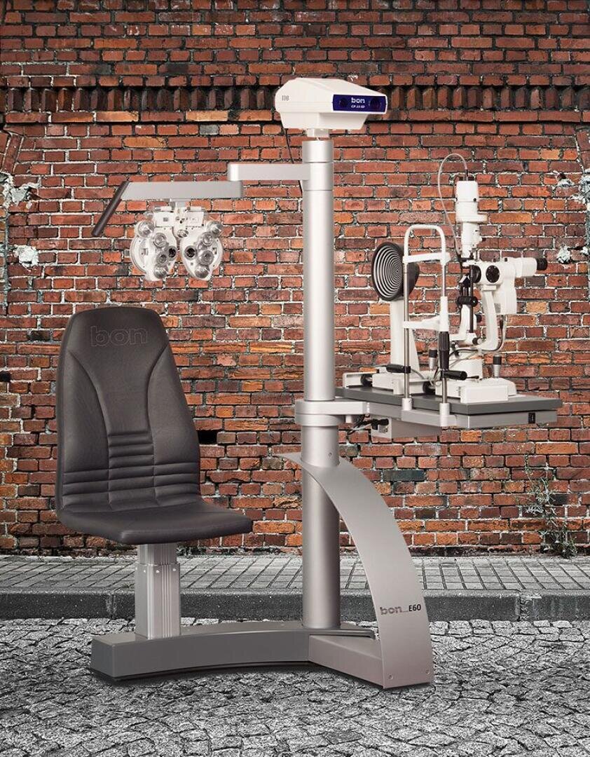 Ein optometrischer Untersuchungsplatz mit einem schwarzen Patientenstuhl, einem Phoropter und einer Spaltlampe, aufgestellt im Freien vor einer verwitterten Backsteinmauer auf einem Kopfsteinpflaster.
