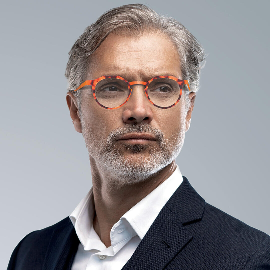 A middle-aged man with gray hair and a beard, wearing a dark suit, white shirt, and round orange-patterned glasses, looks confidently into the distance against a plain gray background.