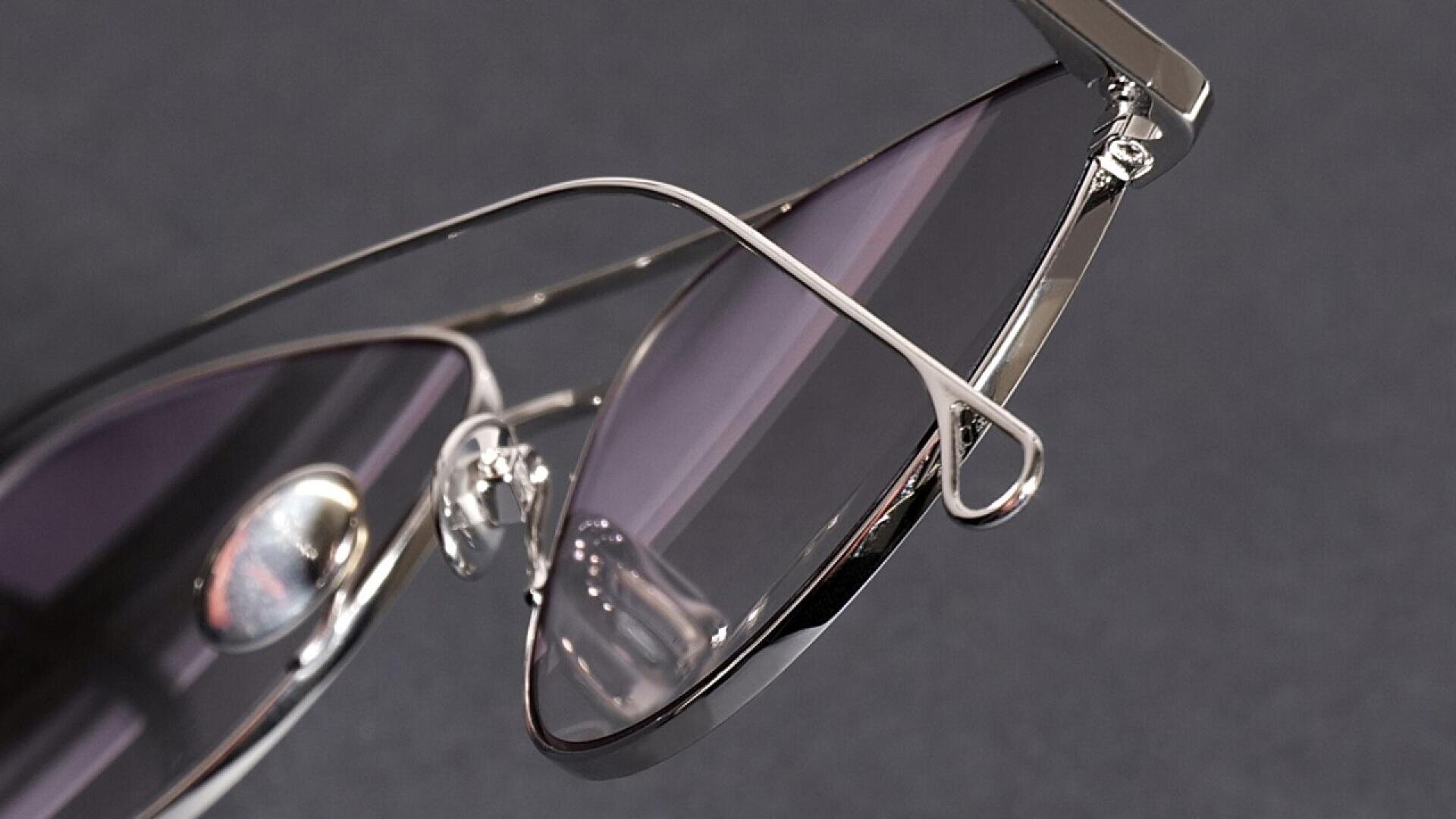 Close-up of stylish sunglasses with thin silver metal frames and purple-tinted lenses, set against a dark background. The focus is on the temples and nose pads.