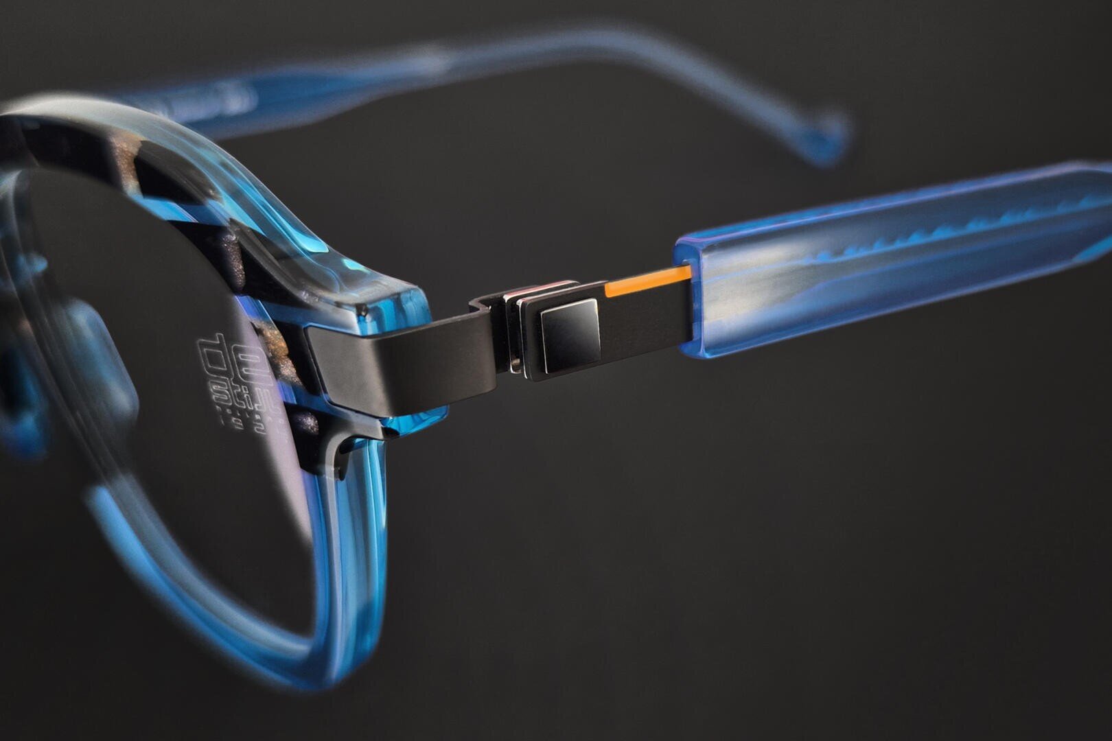 Close-up of a pair of blue, transparent glasses, highlighting the hinge where the frame meets the temple and showing metal and plastic parts against a dark background.