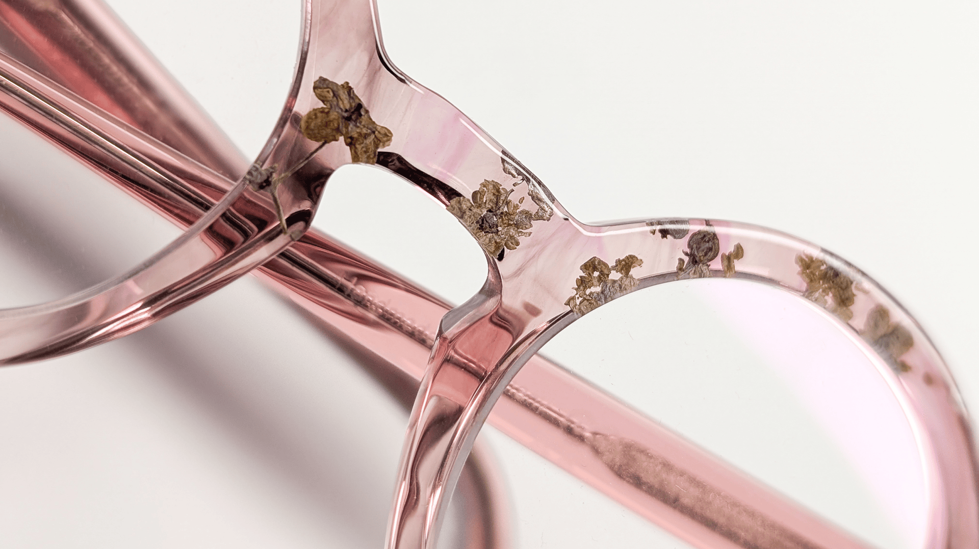 Nahaufnahme einer durchscheinenden rosafarbenen Brille mit kleinen getrockneten Blumen in den Rahmen, vor einem weißen Hintergrund.