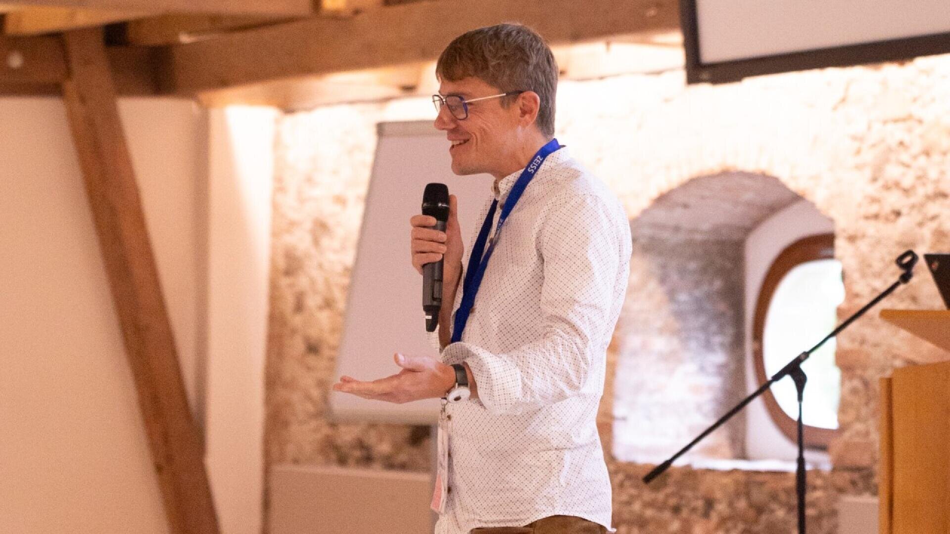 Eine Person mit Brille und weißem Hemd hält ein Mikrofon und spricht vor einem Publikum in einem hellen, rustikalen Raum mit freiliegenden Holzbalken und Steinwänden.