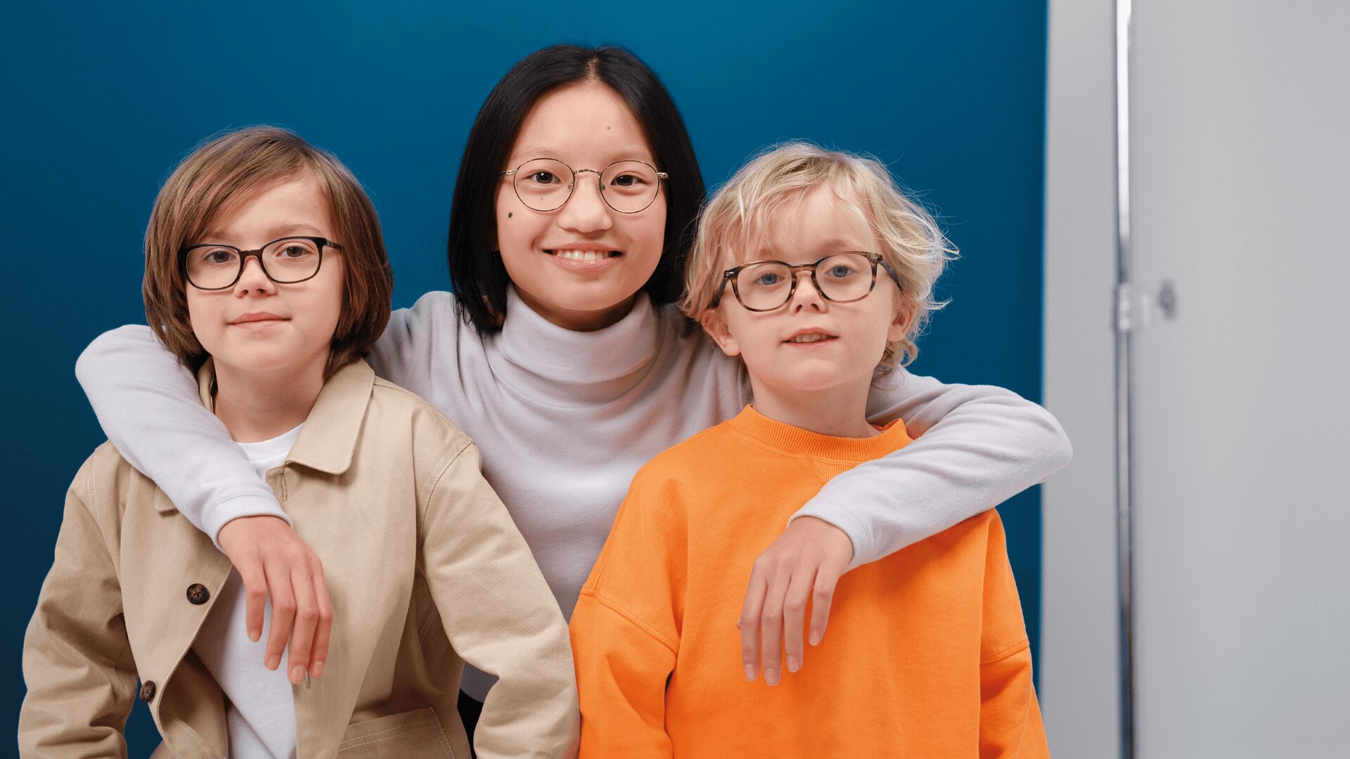 Drei Kinder mit Brillen posieren gemeinsam vor einem blau-weißen Hintergrund. Das Kind in der Mitte, das ein weißes Oberteil trägt, hat seine Arme um die beiden anderen gelegt, die mit beigen und orangefarbenen Oberteilen bekleidet sind. Alle lächeln in die Kamera.