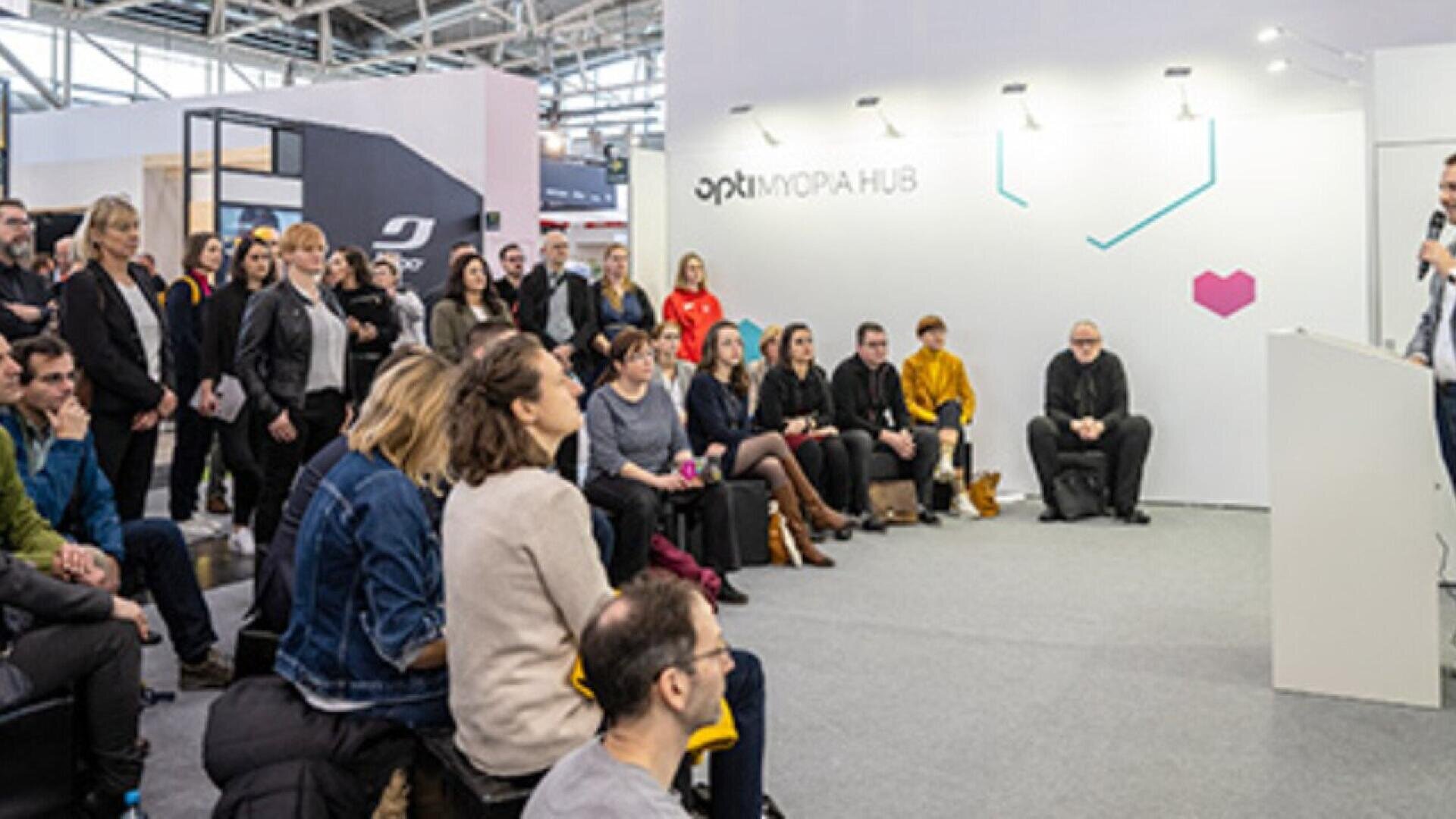 A group of people sits and stands attentively, listening to a speaker at a modern indoor event space with a white wall labeled optinova HUB. The setting appears to be a conference or seminar.