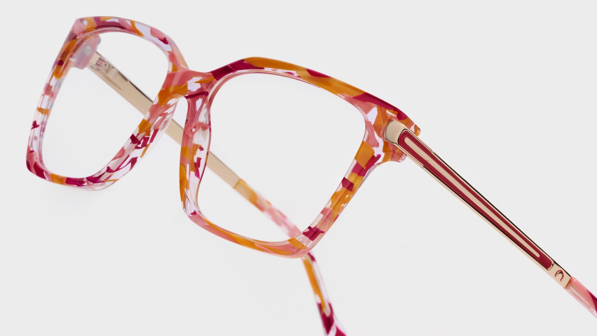 Nahaufnahme einer modischen Brille mit einem auffälligen, mehrfarbigen Rahmen mit rosa, orangefarbenen und weißen Mustern und goldenen Metallbügeln vor einem schlichten weißen Hintergrund.