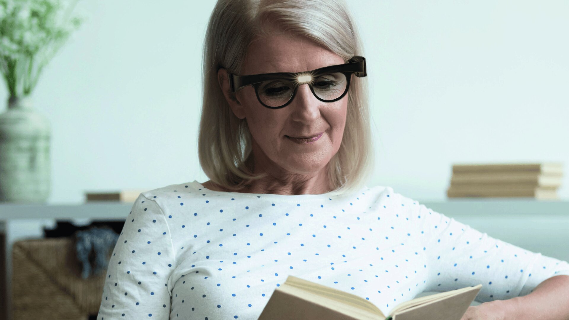 Eine Frau mit grauen Haaren und Brille, die ein weißes Hemd mit blauen Punkten trägt, liest lächelnd ein Buch in einem Innenraum, mit Regalen und einer Blumenvase im Hintergrund.