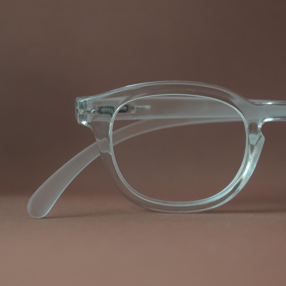 Close-up of a transparent spectacle frame with clear lenses and temples against a plain brown background. Only the left half of the glasses is visible.