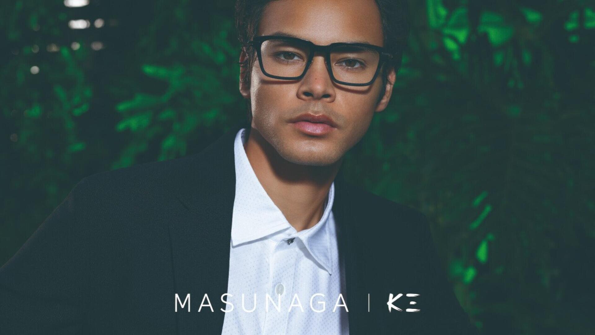 A man wearing black eyeglasses, a white shirt, and a black jacket stands in front of green foliage. The text MASUNAGA | K E appears at the bottom of the image.