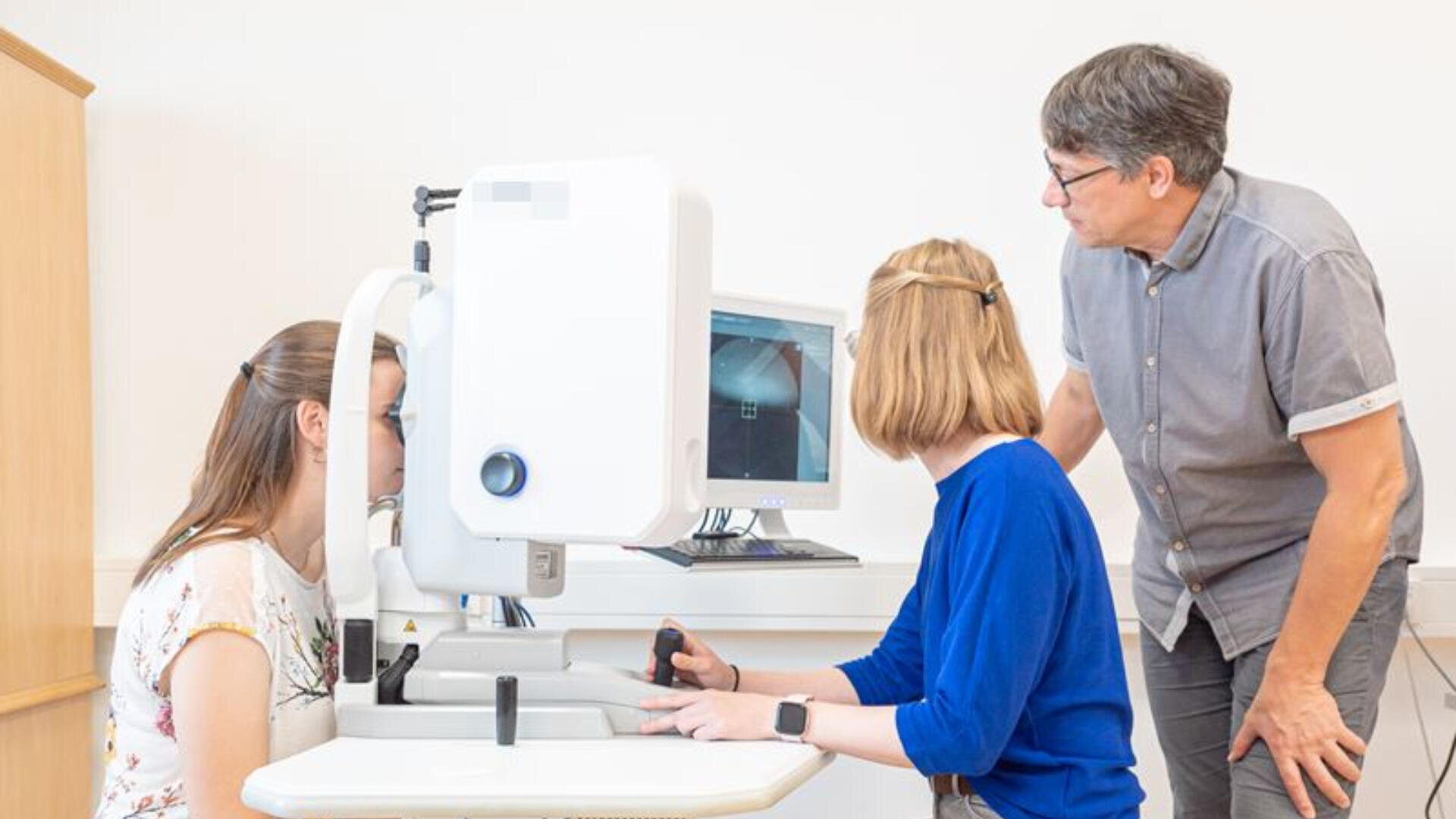 Drei Personen in einer medizinischen Einrichtung verwenden ein Gerät zur Augenuntersuchung. Eine Person wird untersucht, eine andere bedient das Gerät und eine dritte Person beaufsichtigt den Vorgang und schaut dabei auf einen Computermonitor, auf dem die Testergebnisse angezeigt werden.