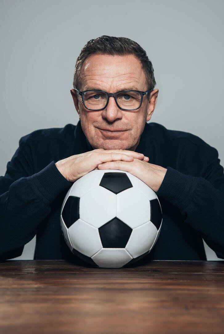 Ein Mann mittleren Alters mit Brille stützt seine Hände und sein Kinn auf einen Fußball, sitzt an einem Holztisch und blickt direkt in die Kamera.